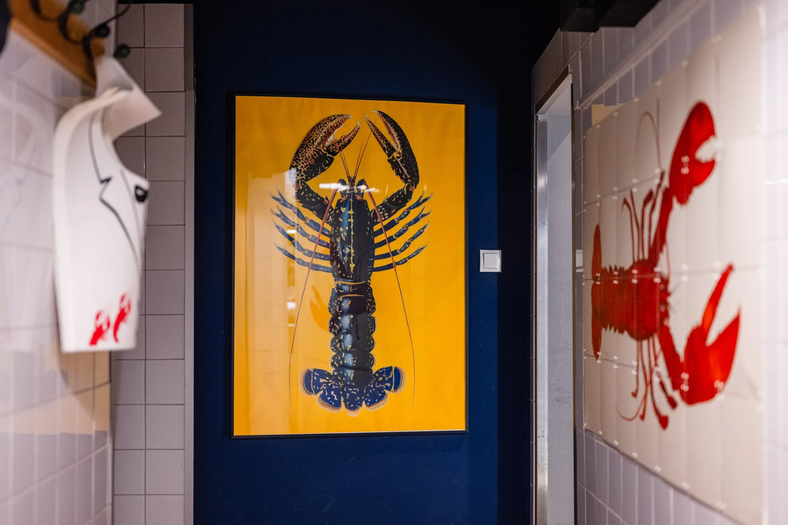 Schmaler Flur mit gelbem Hummer-Poster in schwarzem Rahmen an blauer Wand, rechts ein rotes Hummerbild auf weißen Fliesen.