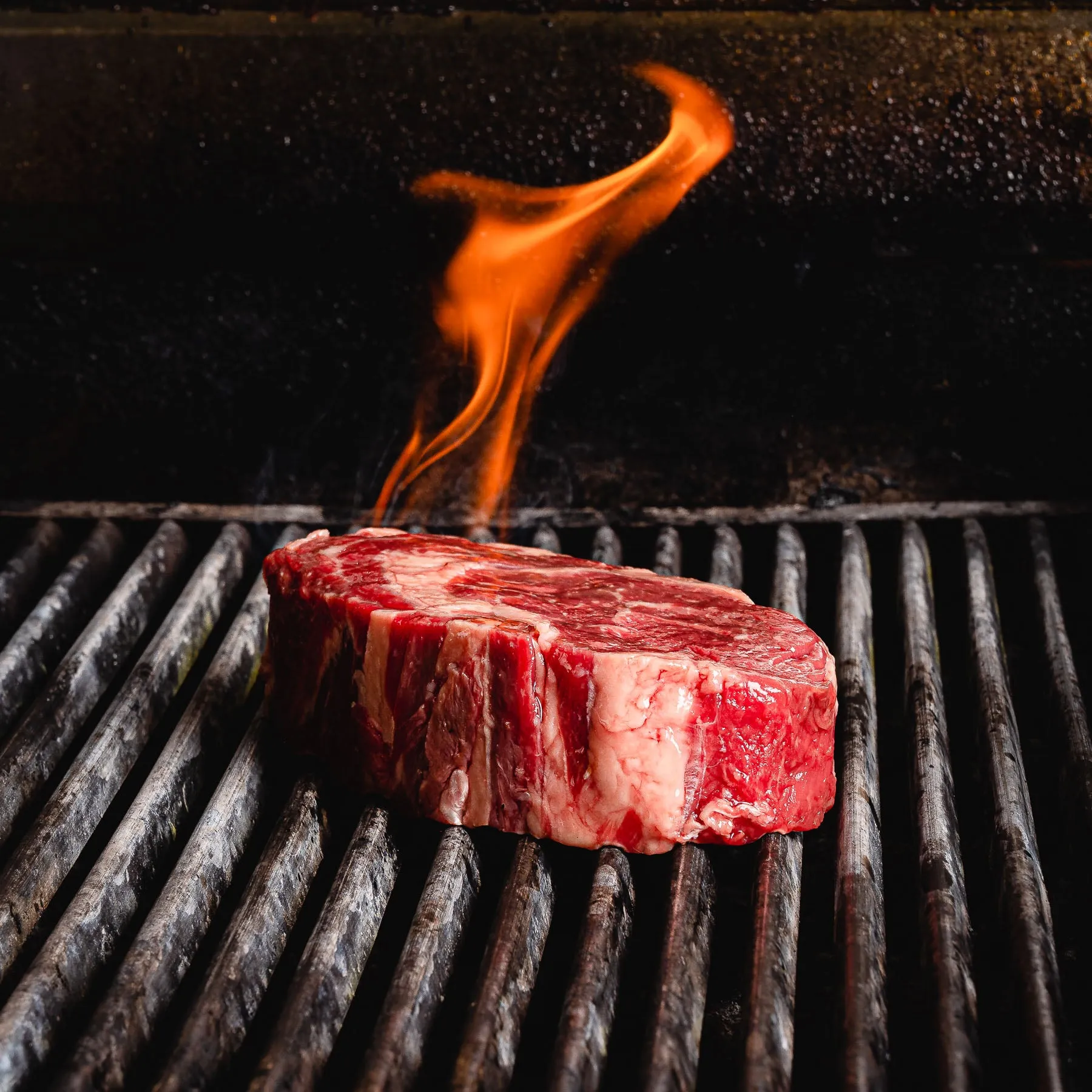 Rohes, stark marmoriertes Rib-Eye-Steak auf dem Rost eines Grills, über dem eine einzelne Flamme aufsteigt.