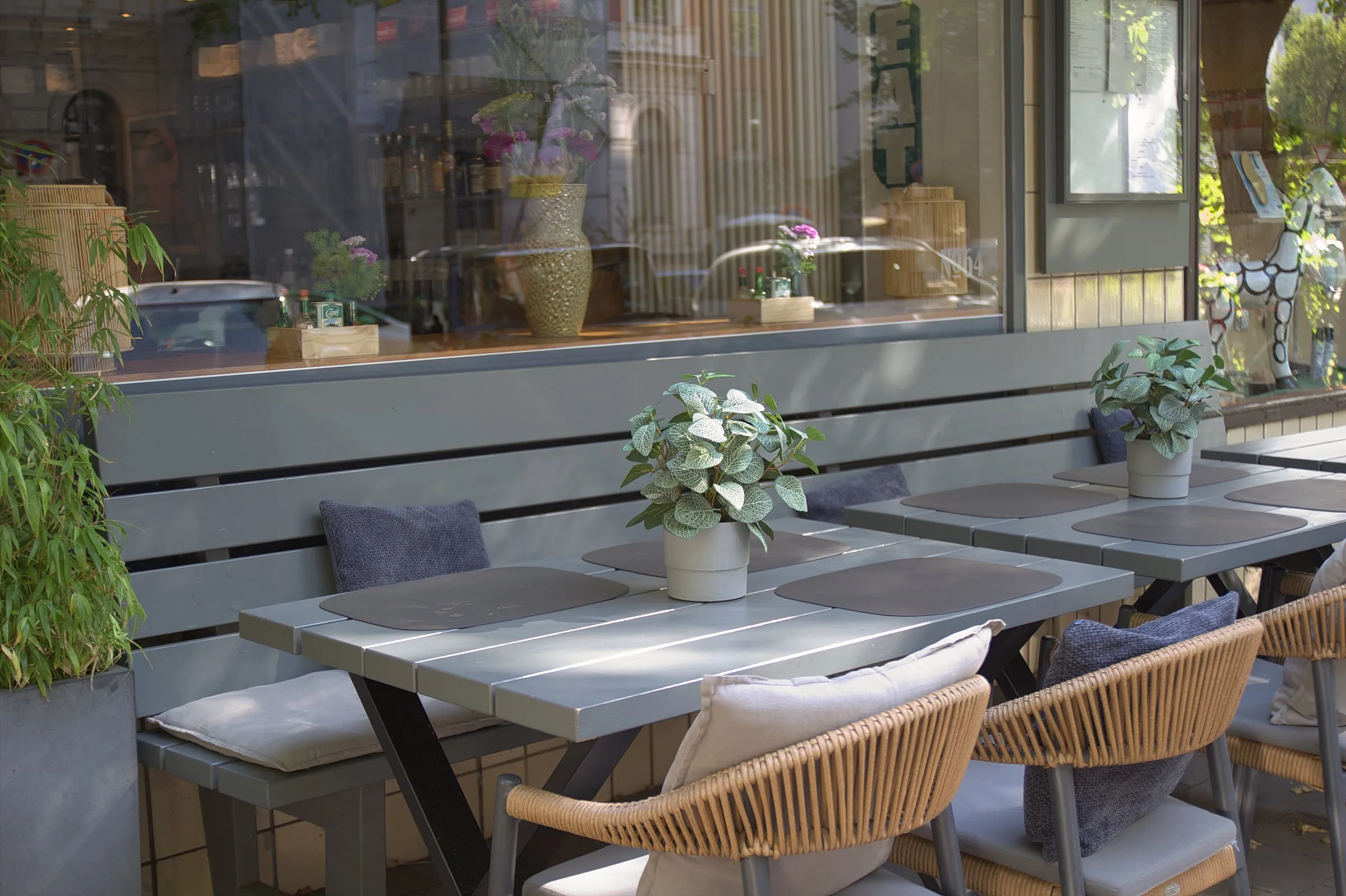 Nahaufnahme der Terrasse mit grauer Bank, Kissen, Tischen mit Pflanzen und Rattanstühlen vor dem Schaufenster.