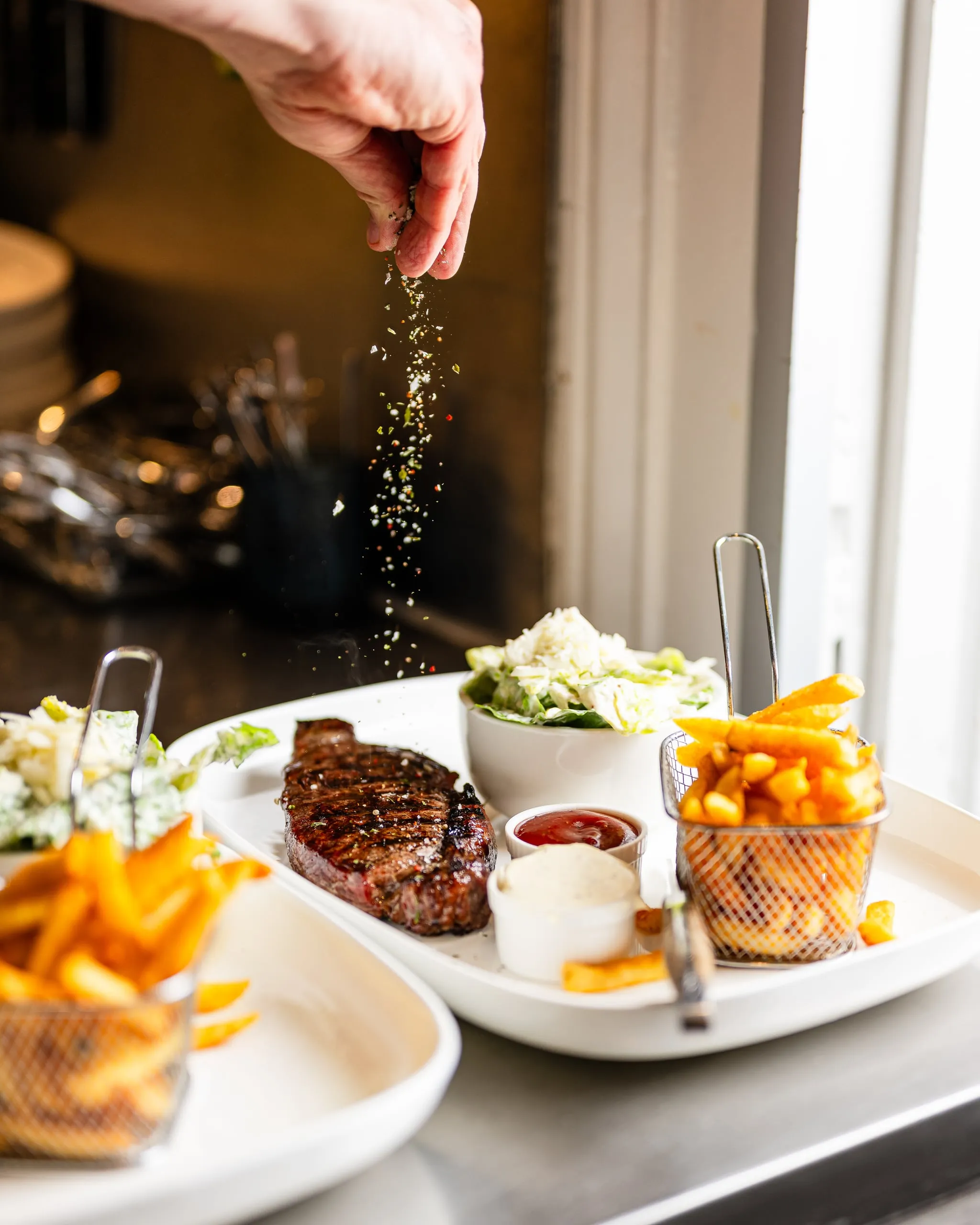 Eine Hand streut Gewürzsalz über ein gegrilltes Steak mit Pommes im Drahtkorb, Salat und Dips auf einer weißen Platte in der Küche.