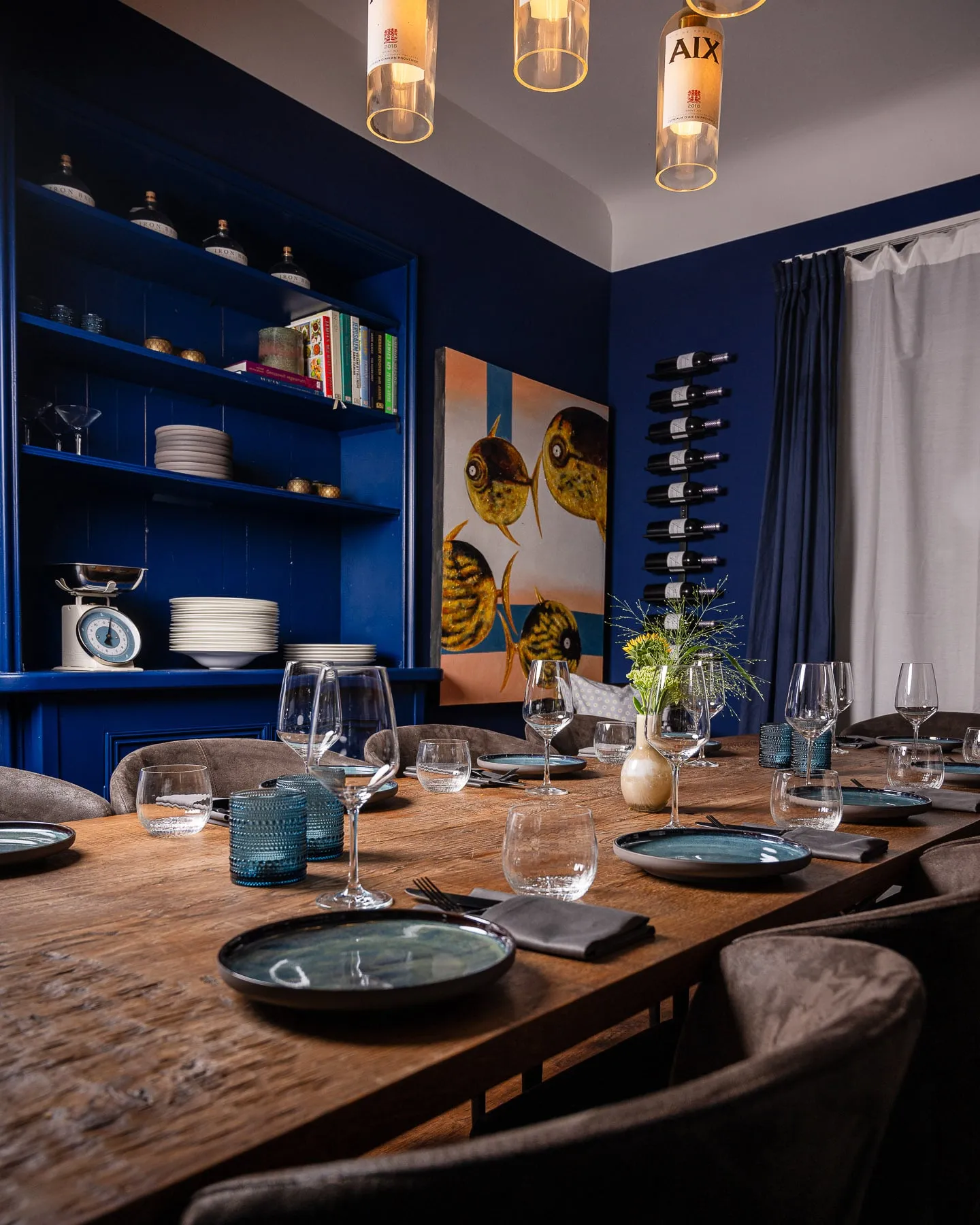 Blaues Esszimmer mit langer Holztafel mit blauen Tellern und Gläsern, links ein Regal mit Büchern und Gin-Flaschen, in der Mitte ein Gemälde mit gelben Fischen, darüber Pendelleuchten aus Weinflaschen.