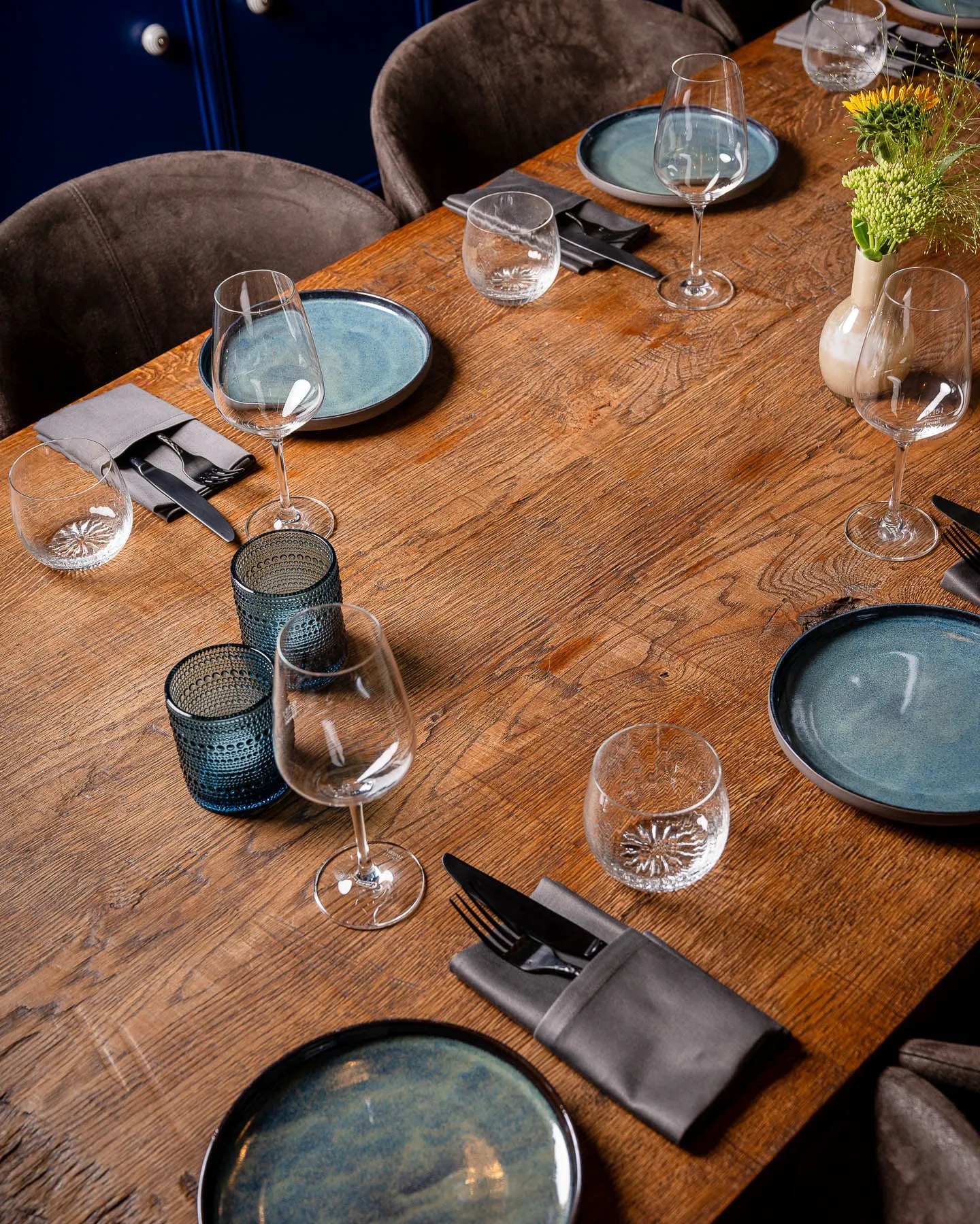 Holztafel von schräg oben mit blauen Tellern, Gläsern, blauen Teelichtgläsern und grauen Servietten, rechts ein Blumenstrauß in einer Vase.