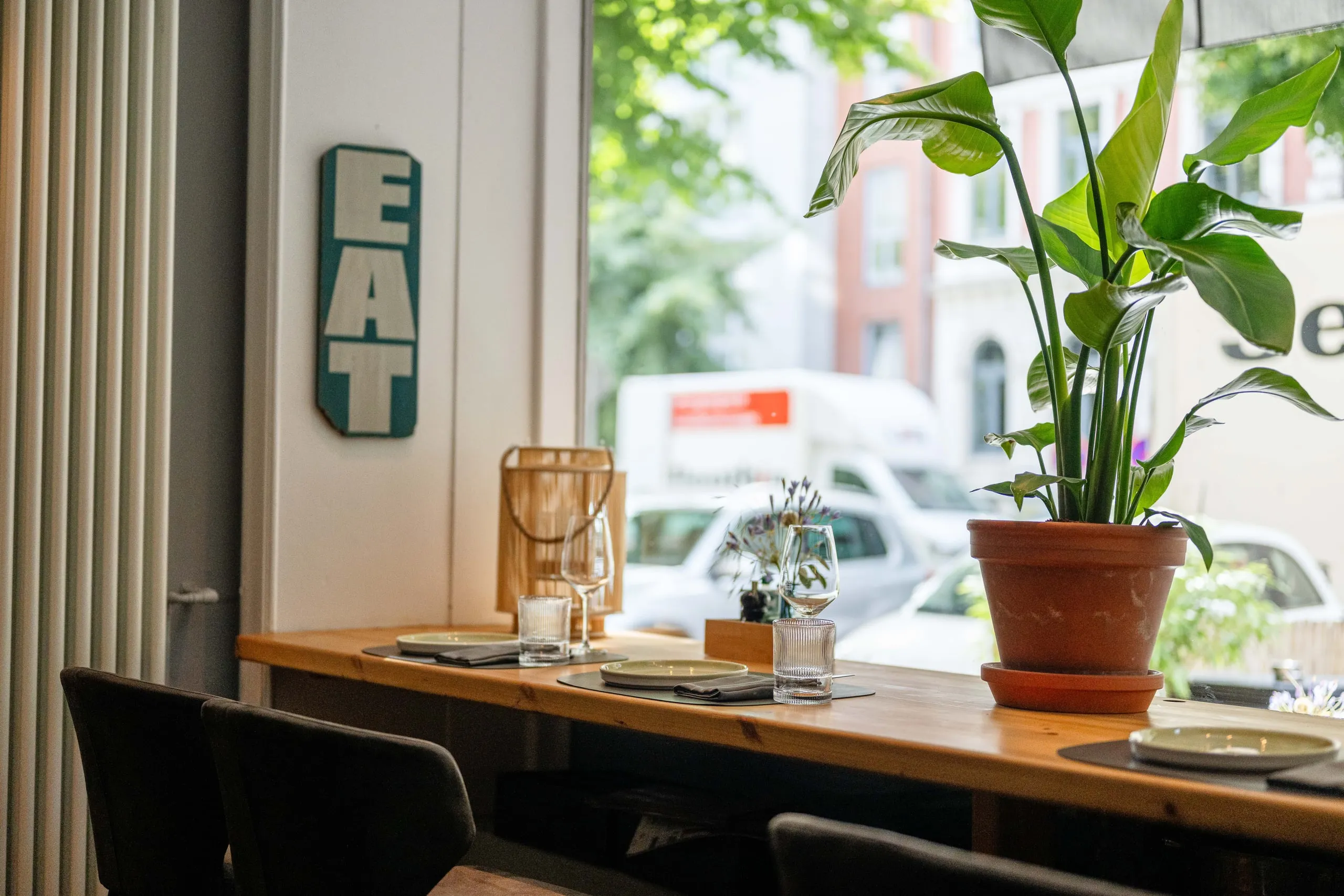 Fensterplatz mit zwei gedeckten Plätzen auf einer Holzplatte, links ein türkisfarbenes EAT-Schild, rechts eine große Zimmerpflanze und Blick auf die Straße.