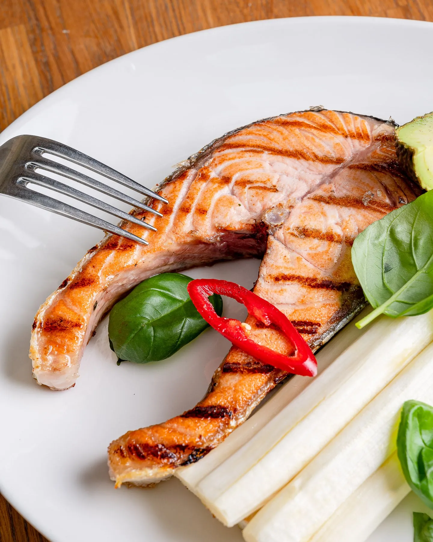 Gegrilltes Lachssteak mit Grillstreifen, weißem Spargel, Basilikum und einer roten Chilischeibe auf einem weißen Teller, links eine Gabel.