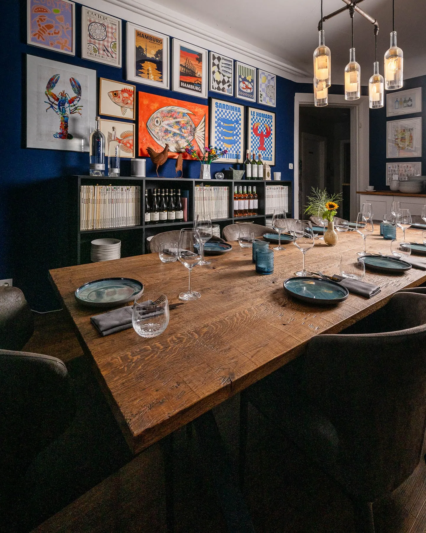 Lange Holztafel im blauen Esszimmer, im Hintergrund eine Wand voller Poster und Bilder, darüber Pendelleuchten aus Weinflaschen.