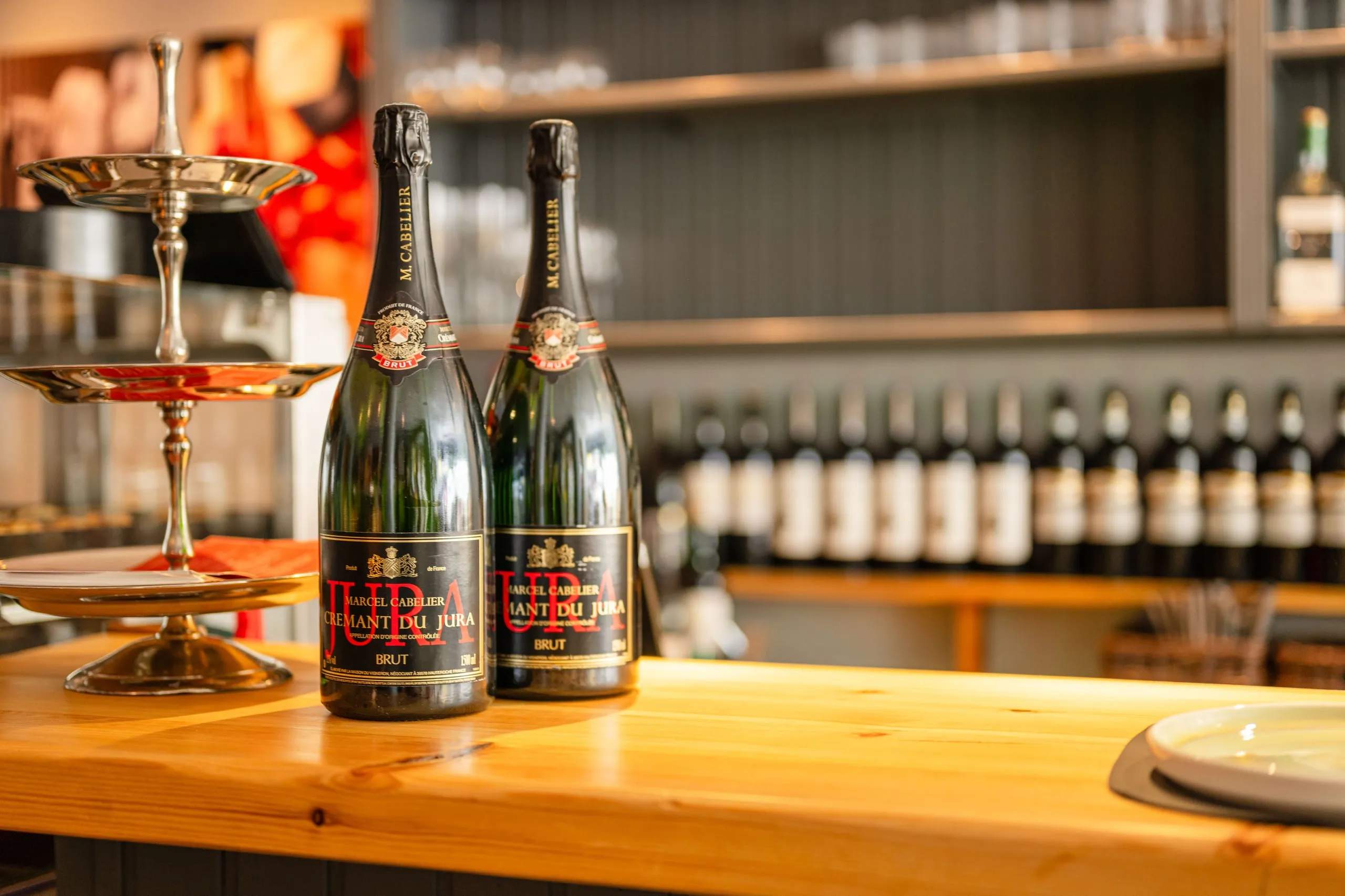 Zwei Flaschen Crémant du Jura Brut auf dem hellen Holztresen, links eine silberne Etagere, im Hintergrund unscharf die Weinflaschen.