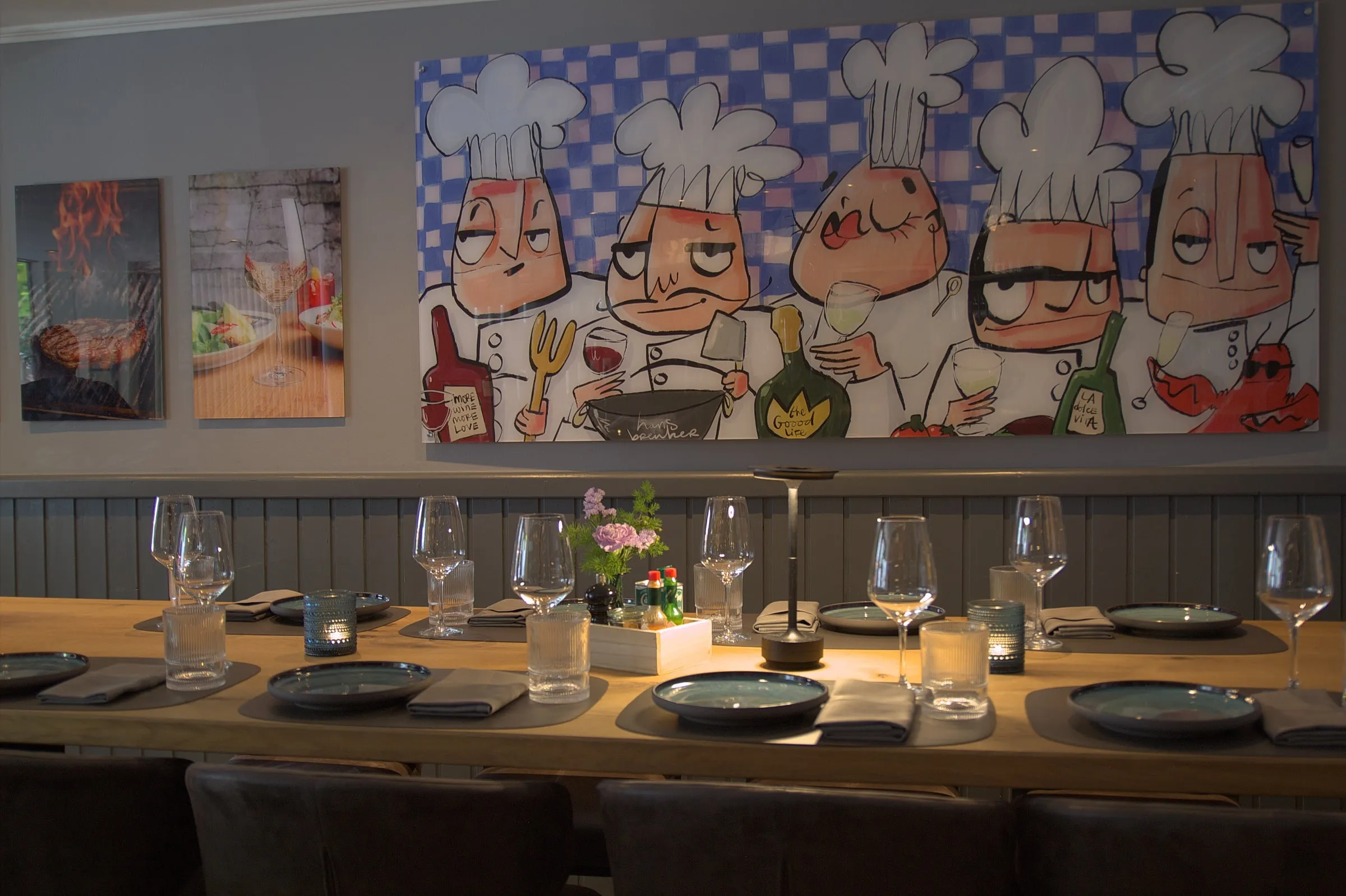 Lange gedeckte Holztafel vor einer grauen Wand, an der ein großes buntes Gemälde mit fünf Köchen hängt, links daneben zwei Fotodrucke.