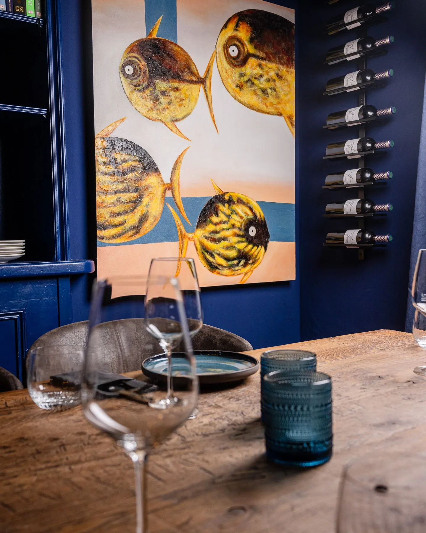 Gemälde mit gelben Fischen an blauer Wand neben einem Weinflaschenhalter, davor die gedeckte Holztafel mit Weinglas und blauen Gläsern.