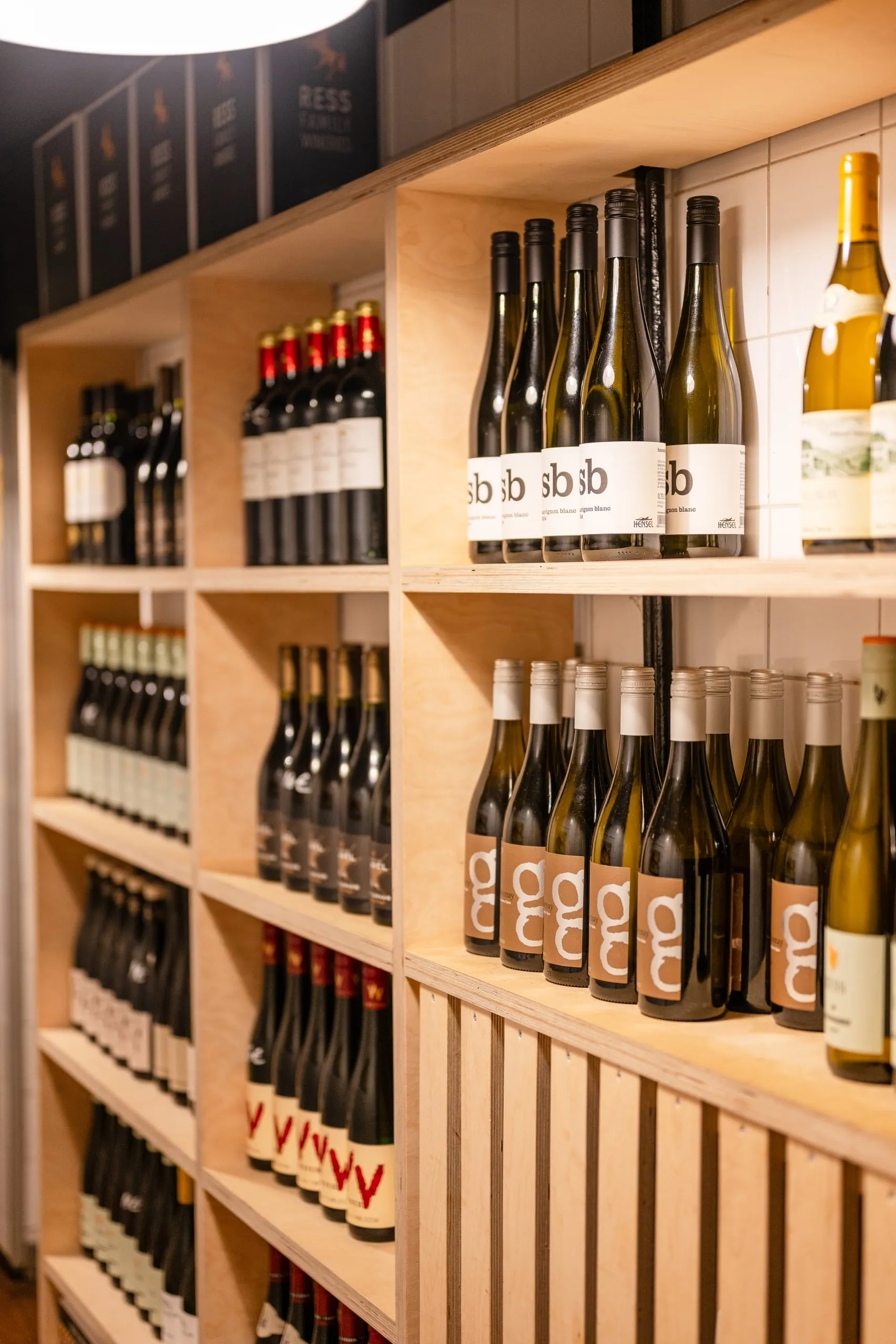 Hohes helles Holzregal mit Rot-, Weiß- und Roséweinflaschen in Reihen, oben dunkle Kartons einer Weinkellerei.