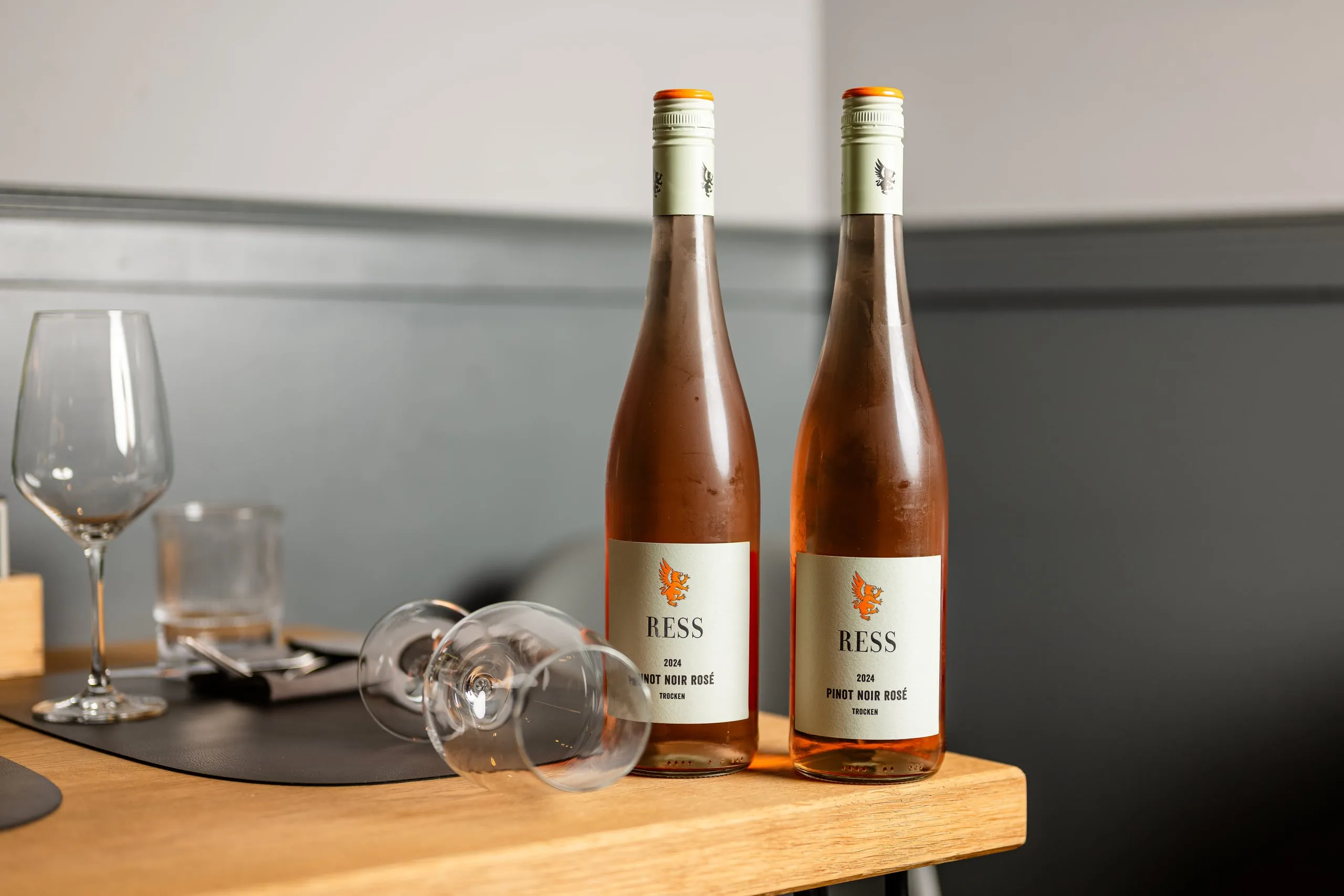 Zwei Flaschen Ress Pinot Noir Rosé 2024 auf einer Holzkante, daneben ein stehendes und ein liegendes Weinglas vor einer grauen Wand.