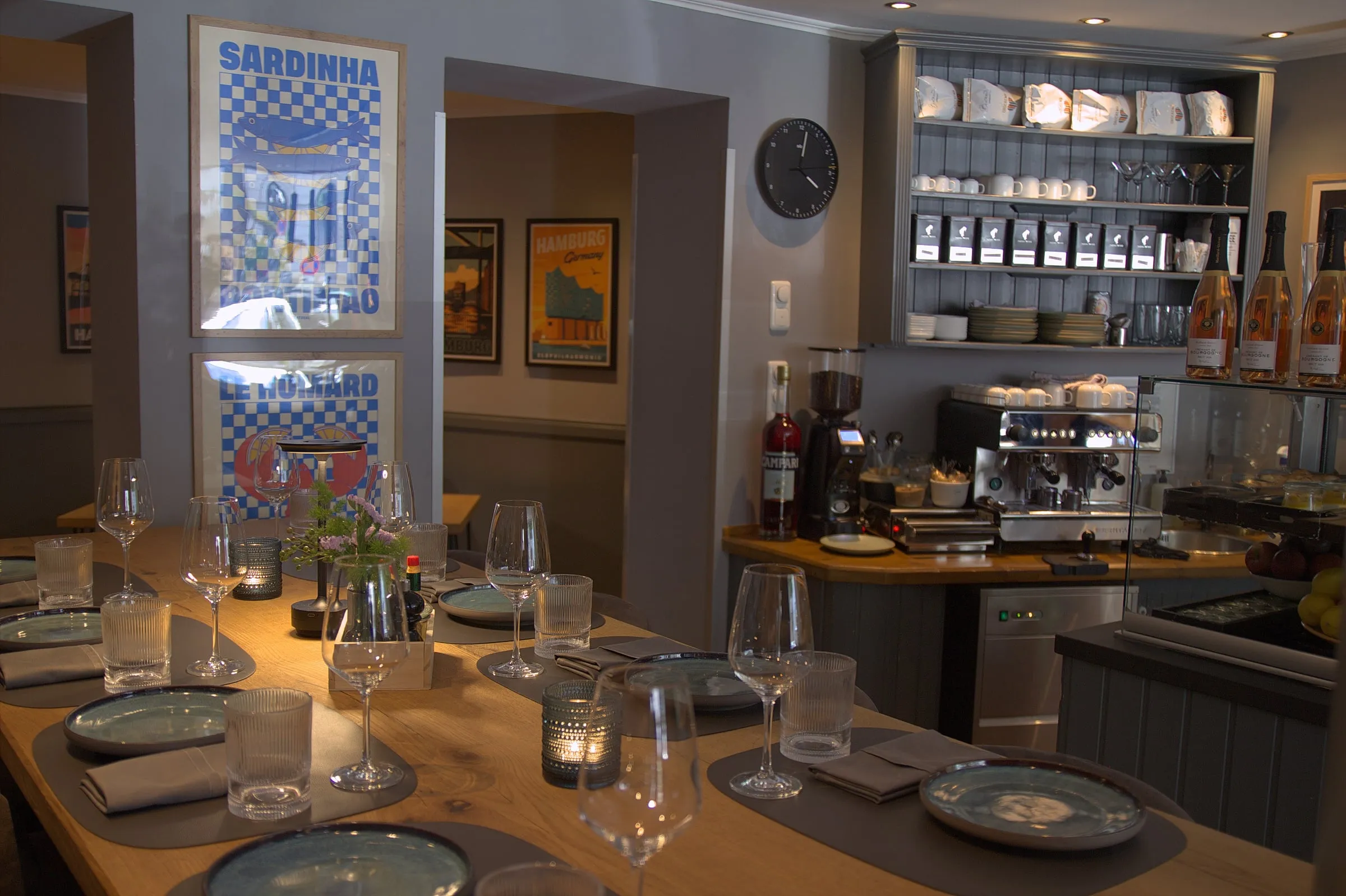 Blick durch den Gastraum über den gedeckten Holztisch zur Bar mit Espressomaschine und Regalen, links die Poster „Sardinha“ und „Le Homard“ und ein Durchgang.