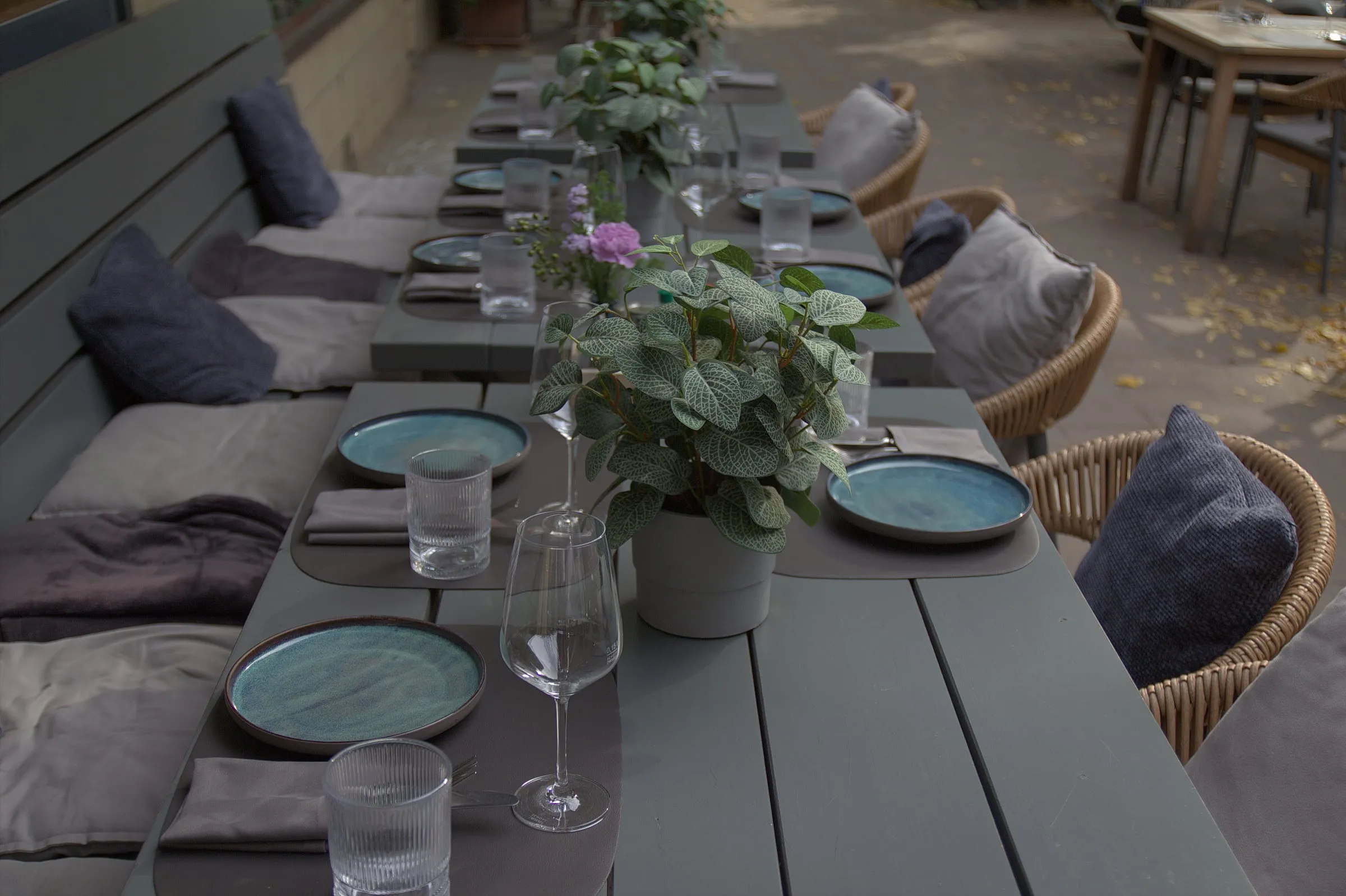 Lange Terrassenbank mit gedeckten Plätzen, blauen Tellern, Gläsern und Pflanzen, dazu Rattanstühle.
