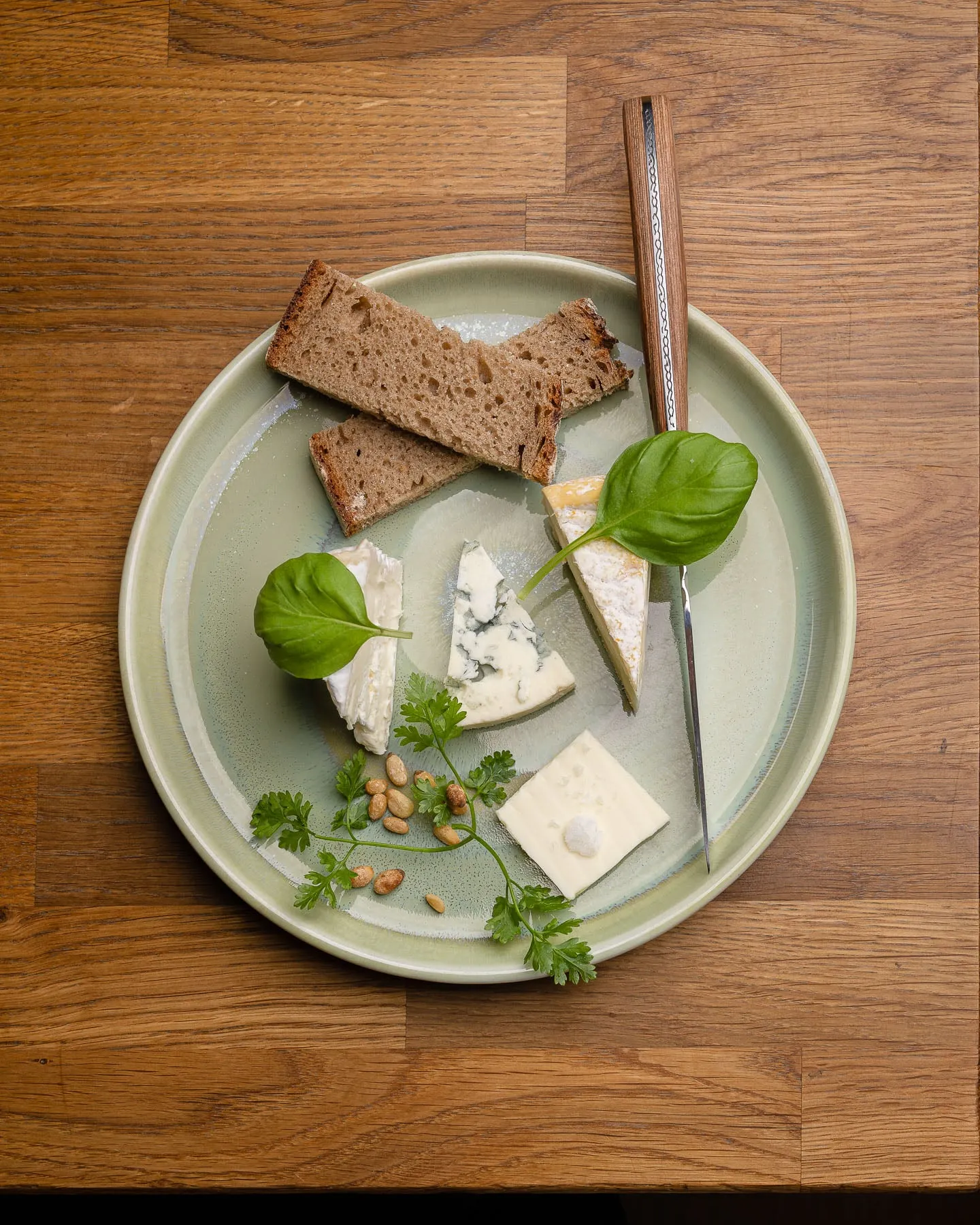 Grüner Teller von oben mit Roggenbrotscheiben, vier Käsesorten, Basilikum, Kerbel und Pinienkernen, daneben ein Messer mit Holzgriff, auf einem Holztisch.