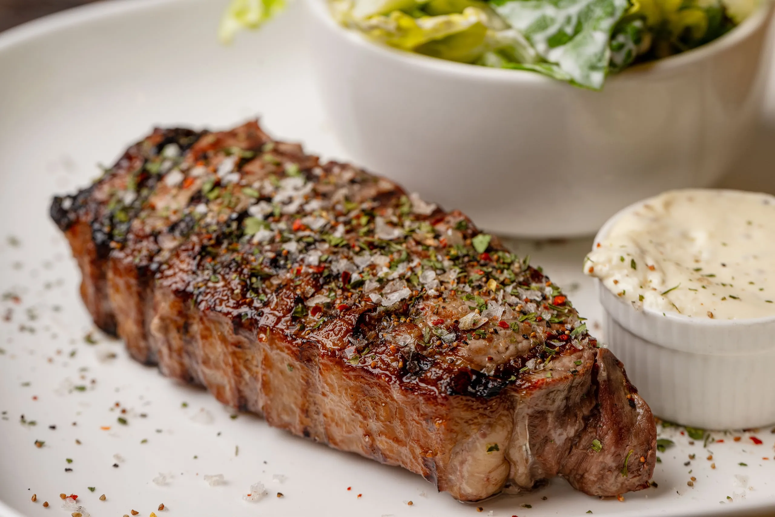 Nahaufnahme eines gegrillten Rumpsteaks mit Salz, Pfeffer und Kräutern auf einem weißen Teller, daneben Kräuterbutter und eine Schale Salat.