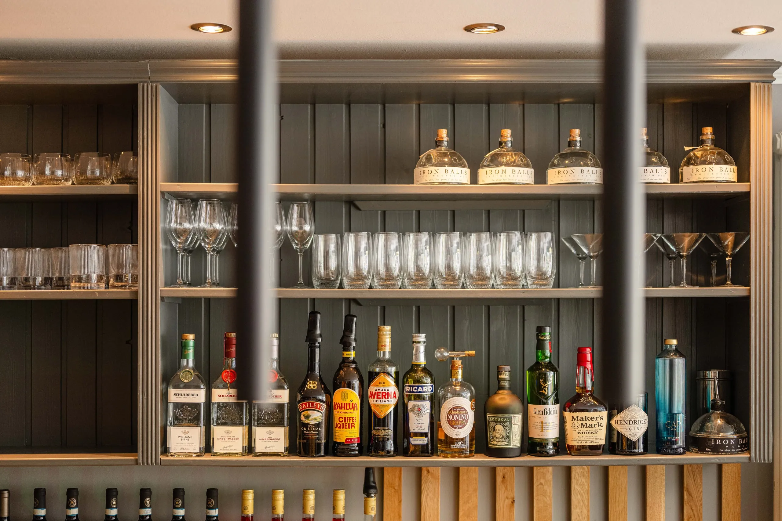 Graues Holzregal der Bar mit Gin-, Likör- und Whiskyflaschen, Weingläsern und Martinischalen, im Vordergrund unscharf zwei schwarze Pendelleuchten.