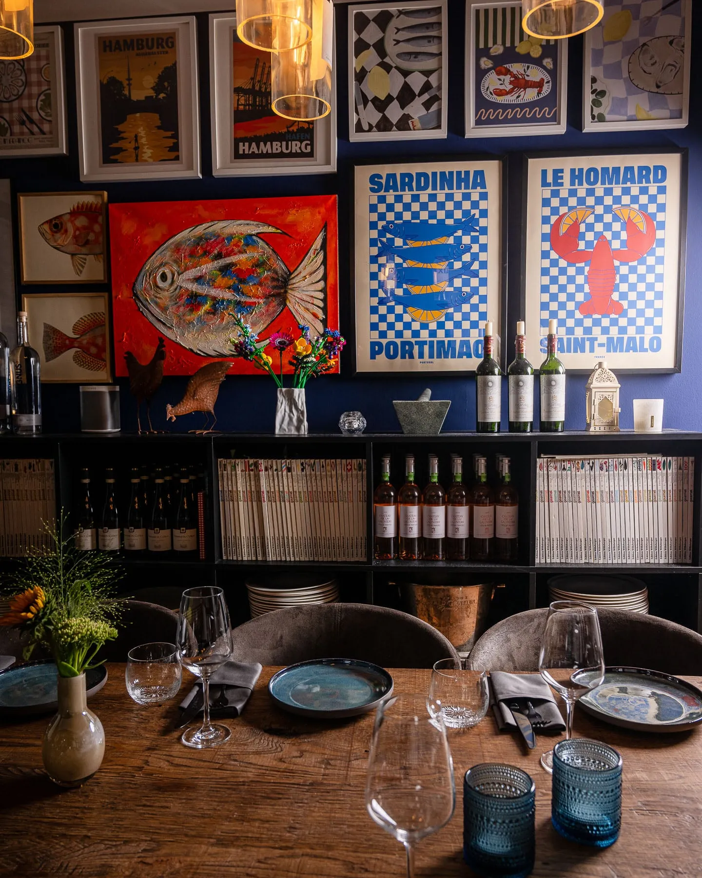 Bilderwand des blauen Esszimmers mit den Postern „Sardinha“ und „Le Homard“, einem roten Fischgemälde und Hamburg-Plakaten, darunter ein Regal mit Weinflaschen und Büchern.