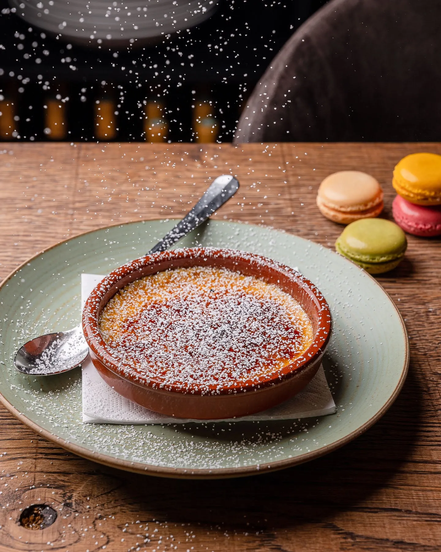 Crème brûlée in einer roten Tonschale mit Puderzucker auf einem grünen Teller, daneben bunte Macarons auf einem Holztisch.