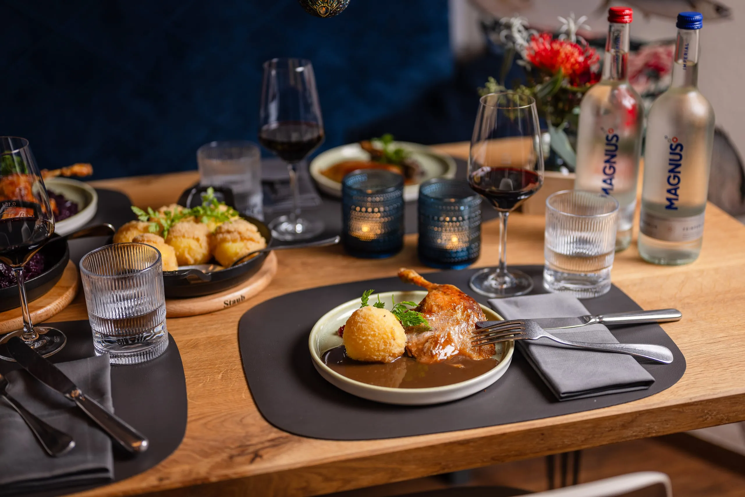 Gedeckter Holztisch mit einem Teller Keule, Knödel und dunkler Soße, daneben Rotweingläser, blaue Windlichter, Magnus-Wasserflaschen und ein Blumenstrauß.