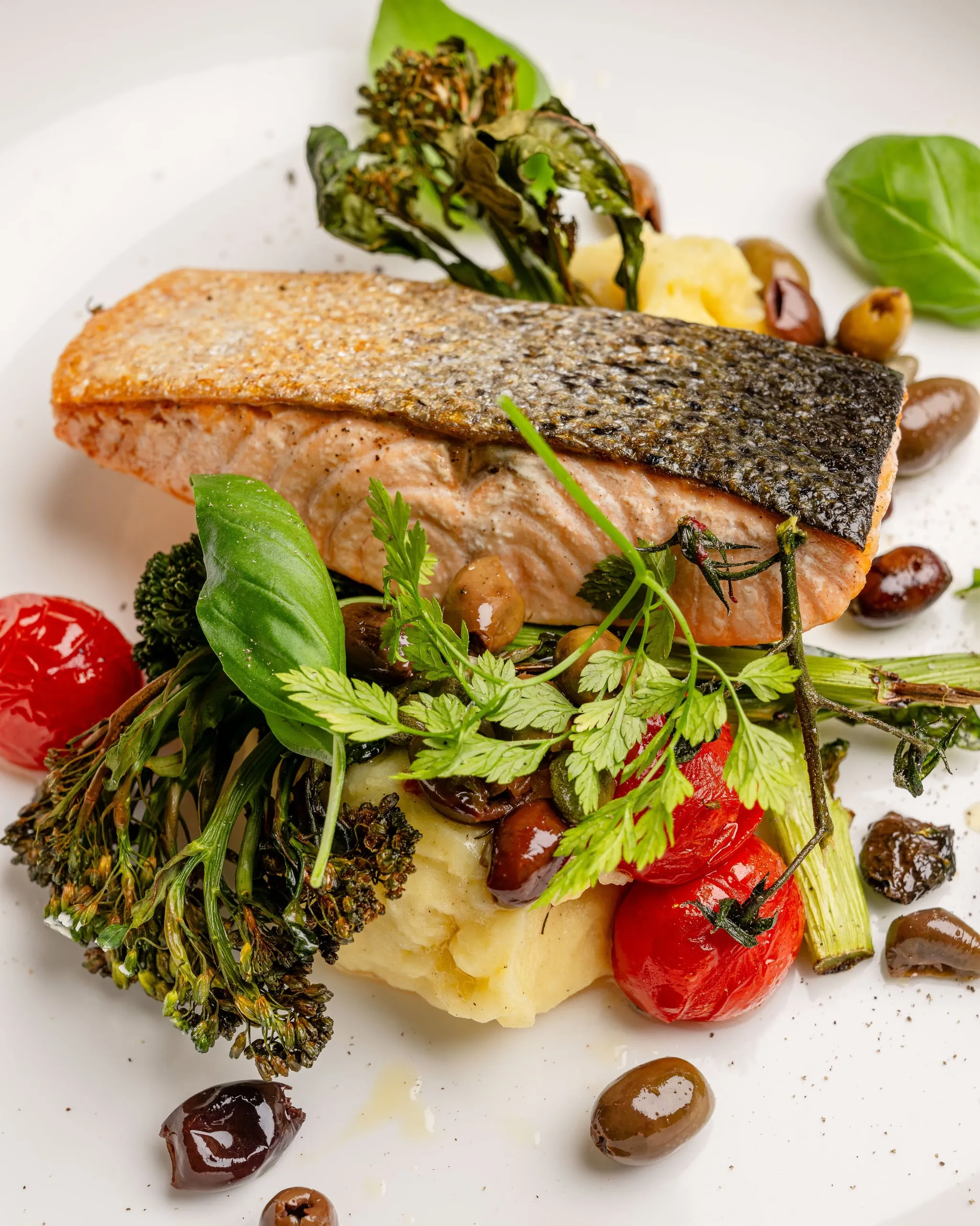 Gebratenes Lachsfilet mit knuspriger Haut auf Kartoffelpüree, umgeben von Brokkoli, Oliven, Kirschtomaten, Kerbel und Basilikum auf einem weißen Teller.