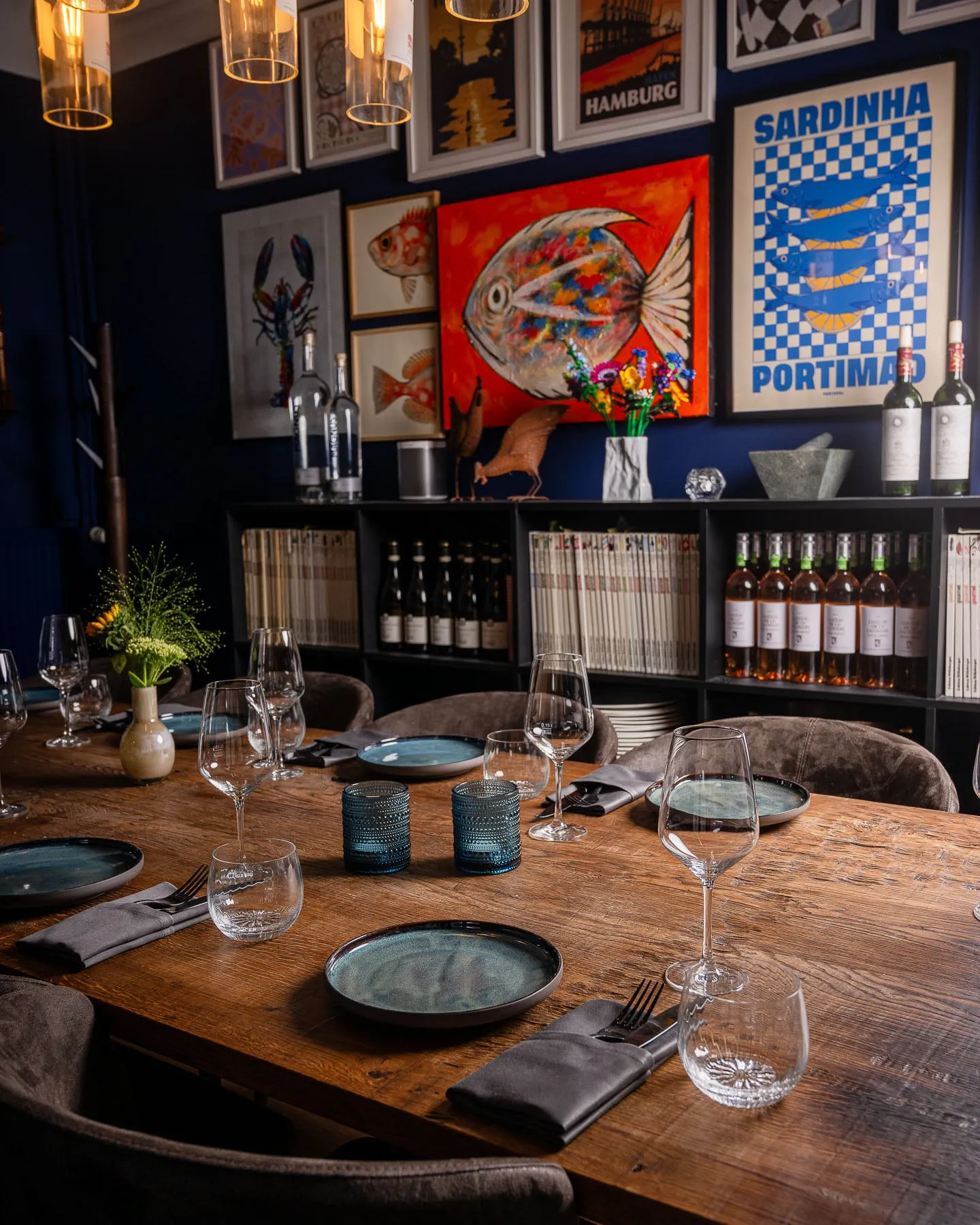 Gedeckte Holztafel im blauen Esszimmer vor einer Bilderwand mit Postern, einem roten Fischgemälde und einem Regal mit Weinflaschen und Büchern.