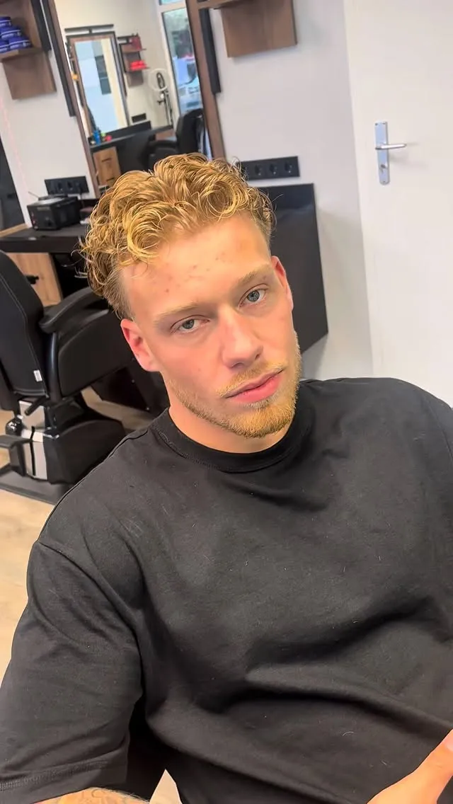 Junger Mann mit kurzen, blond gefärbten Locken und blondem Dreitagebart am Arbeitsplatz des Salons.