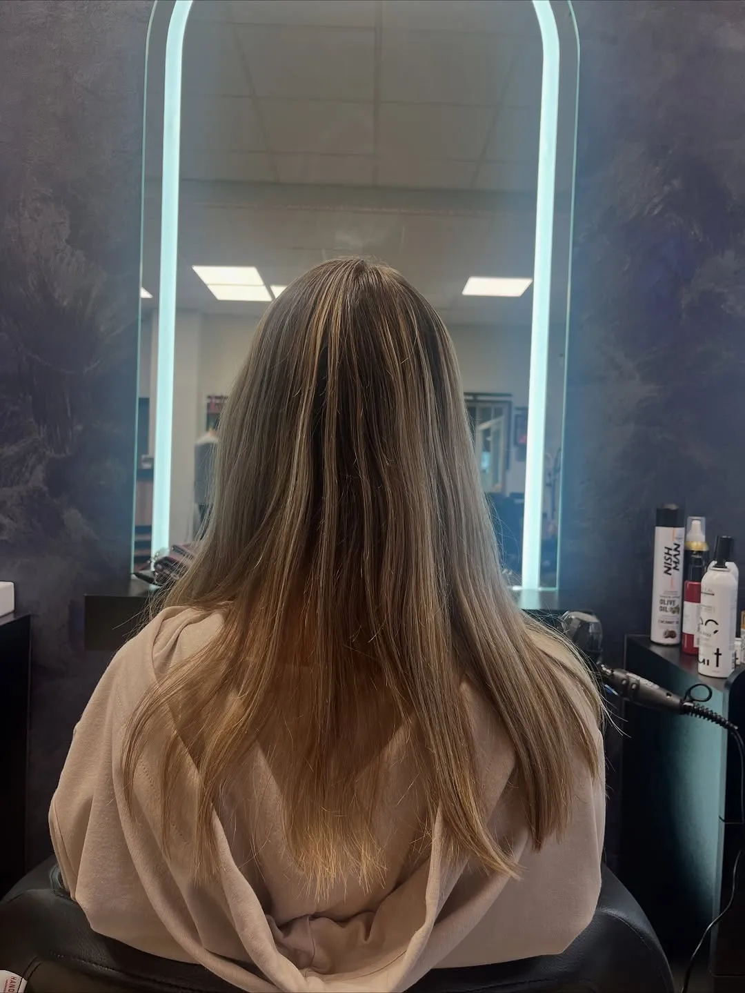 Lange, glatt geföhnte Haare in Dunkelblond mit helleren Spitzen, von hinten vor einem beleuchteten Bogenspiegel.