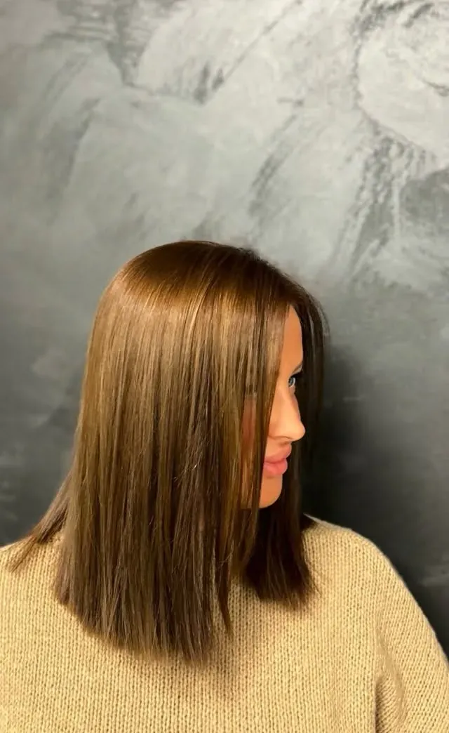 Brünetter Long Bob, stumpf auf Schulterhöhe geschnitten und glatt gestylt, im Profil vor einer grauen Spachtelwand.