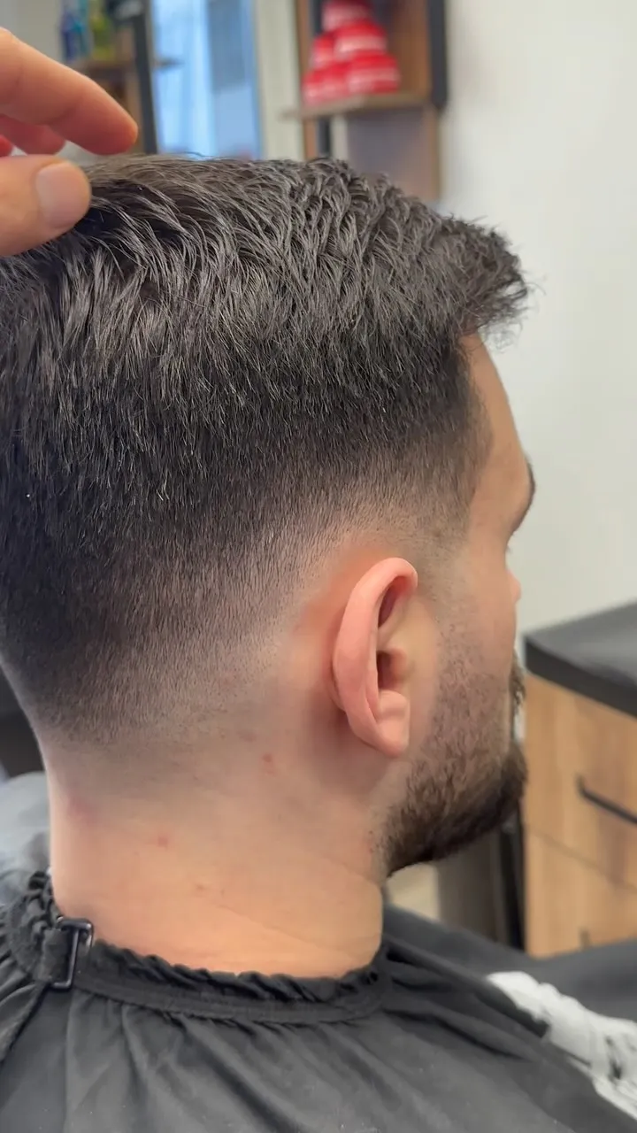 Kurzer dunkler Herrenschnitt mit sauber verlaufendem Fade an Seite und Nacken; eine Hand legt das Deckhaar zur Seite.