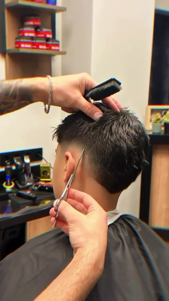 Standbild aus dem Hero-Clip: eine Hand führt Kamm und Schere am Hinterkopf eines Herren-Fades, dahinter das Regal mit Stylingprodukten.