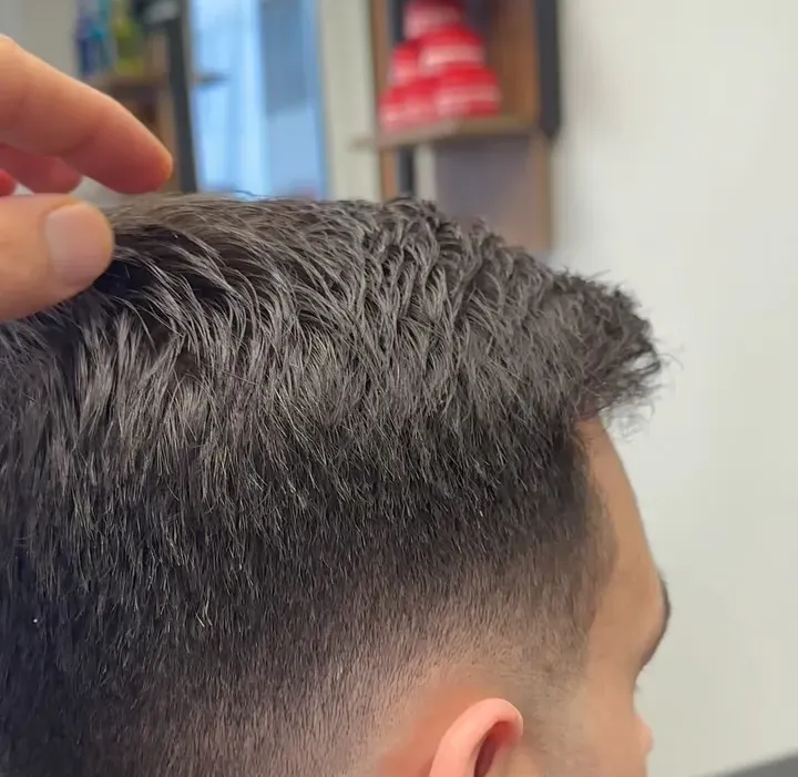 Kurzer dunkler Herrenschnitt mit sauber verlaufendem Fade an Seite und Nacken; eine Hand legt das Deckhaar zur Seite.