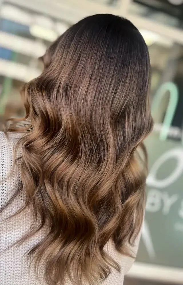 Lange Balayage in Karamellblond, in weiche Wellen gelegt, vor der Fensterfront des Salons; oben rechts steht ein eingeblendetes Schnittprogramm-Wasserzeichen.