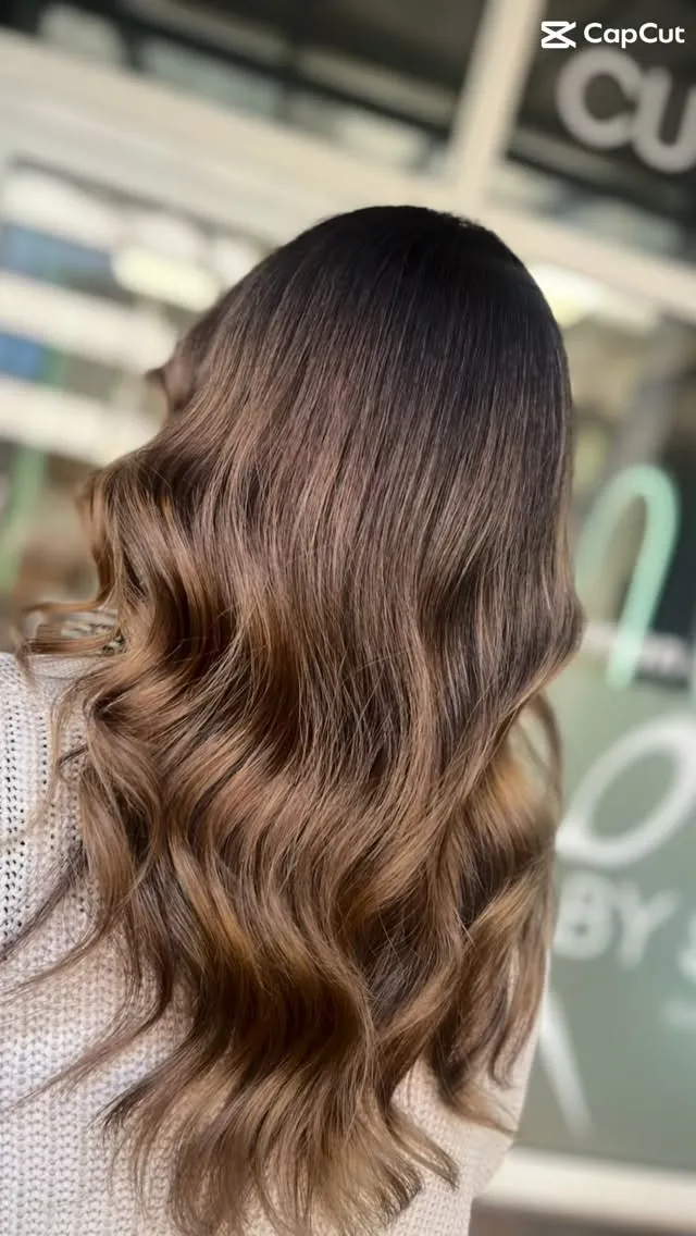 Lange Balayage in Karamellblond, in weiche Wellen gelegt, vor der Fensterfront des Salons; oben rechts steht ein eingeblendetes Schnittprogramm-Wasserzeichen.