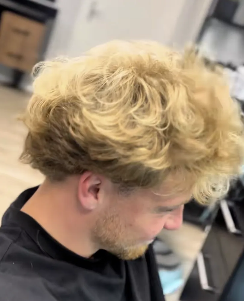Vorher: blondes, ausgewachsenes Haar mit unruhigen, undefinierten Locken; der Kunde trägt den schwarzen Frisierumhang.
