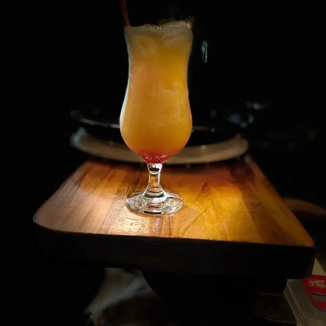 Orangefarbener Cocktail in einem Hurricane-Glas auf einem Holzbrett, im Spotlicht vor dunklem Hintergrund.