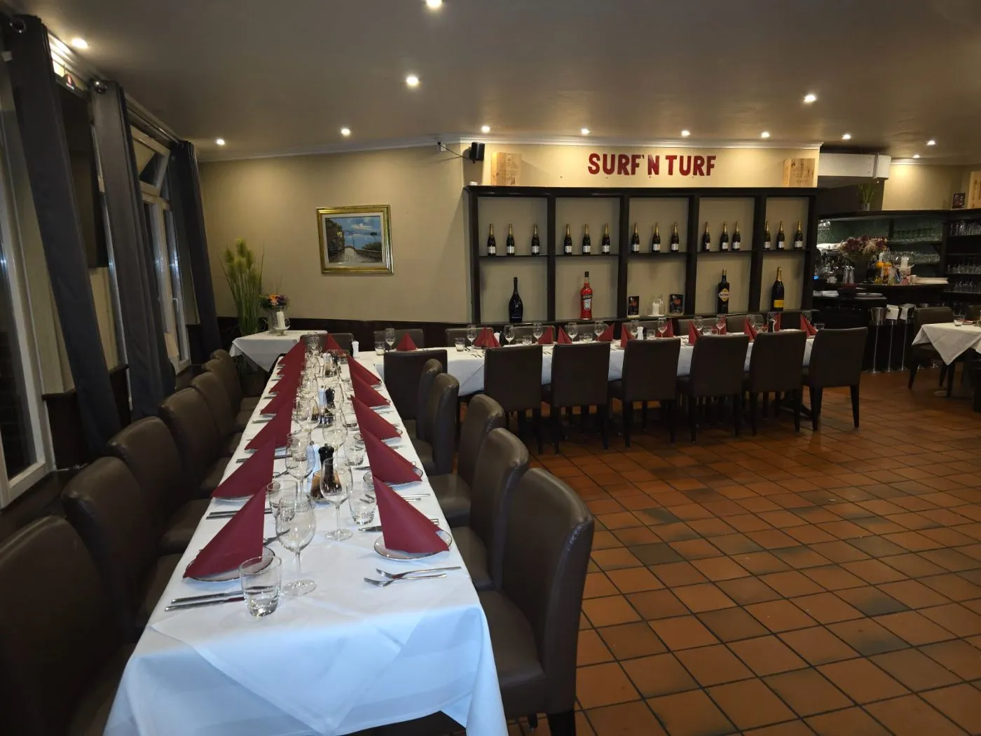 Hinterer Bereich mit einer langen, weiß gedeckten Tafel und roten Servietten, an der Wand ein Weinregal mit dem Schriftzug „Surf N Turf“.