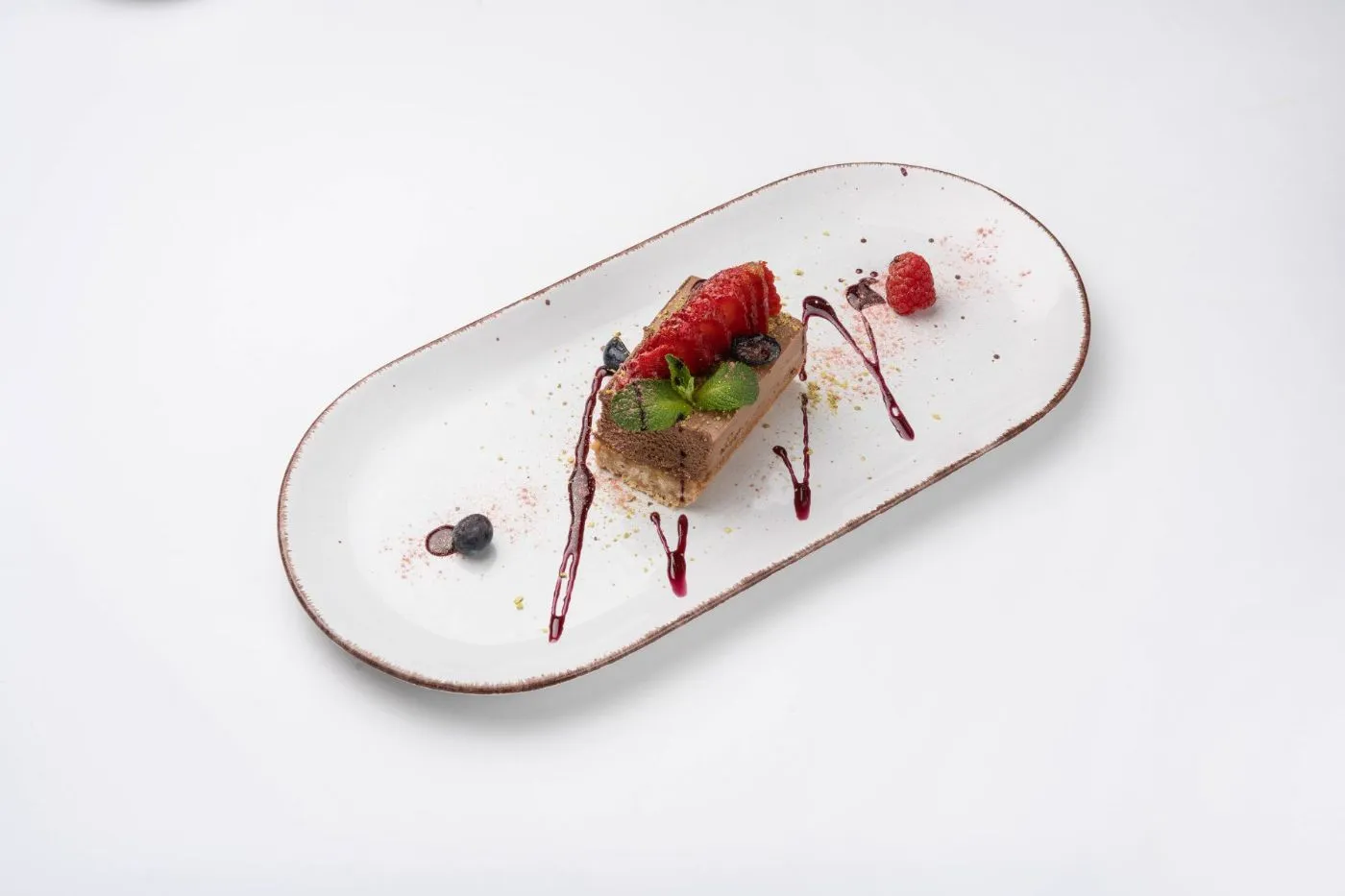 Cheesecake-Schnitte mit frischen Beeren und Fruchtsauce auf einem ovalen weißen Teller vor weißem Grund.