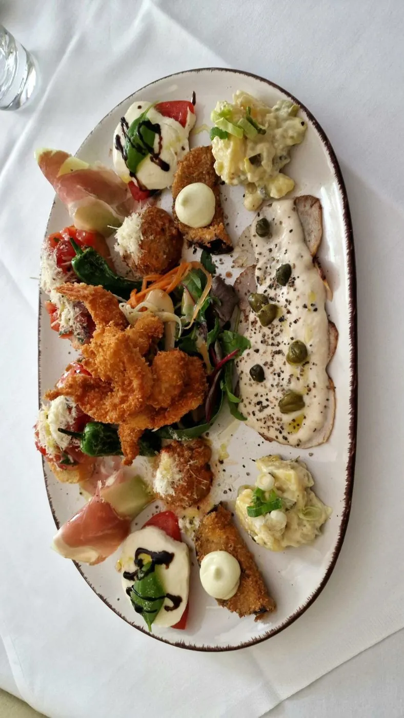 Große Vorspeisenplatte mit Schinken, Fleischbällchen, Antipasti und Dips auf einer länglichen weißen Platte.
