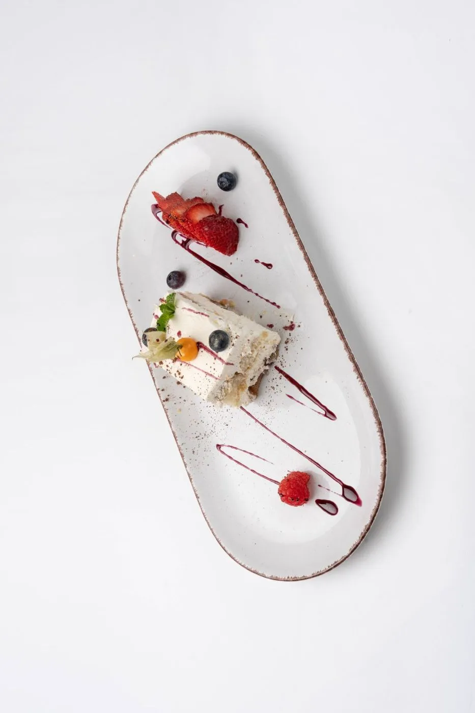 Tiramisu-Schnitte mit Beeren und Fruchtsauce auf einem ovalen weißen Teller, von oben.