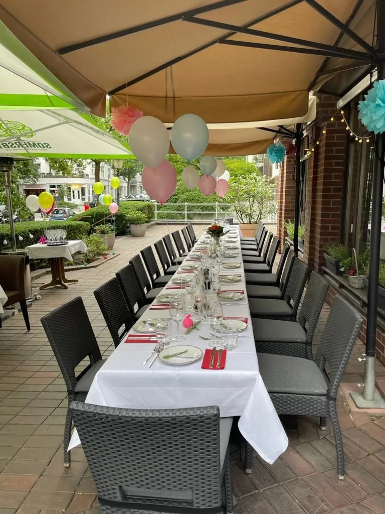 Terrasse unter der Markise mit langer Tafel für eine Feier, Ballons und Lichterkette, Blick auf die Straße.
