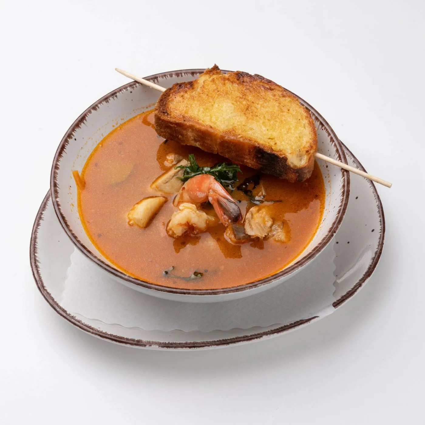 Fischsuppe mit Garnelen in einer weißen Schale, obenauf eine geröstete Brotscheibe am Spieß.