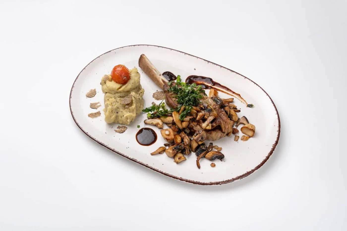 Kalbskarree mit Pilzen, Gemüsepüree und Kirschtomate auf einem ovalen Teller vor weißem Grund, im Querformat.