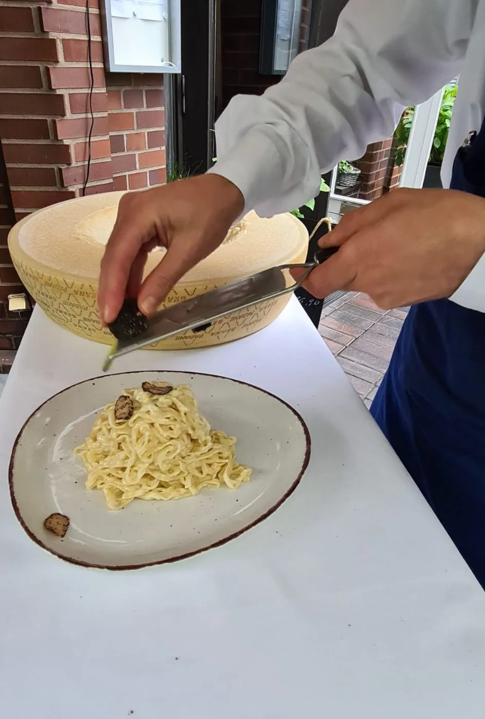 Eine Servicekraft schabt am Tisch Parmesan aus einem Käselaib, davor ein Teller Tagliolini.