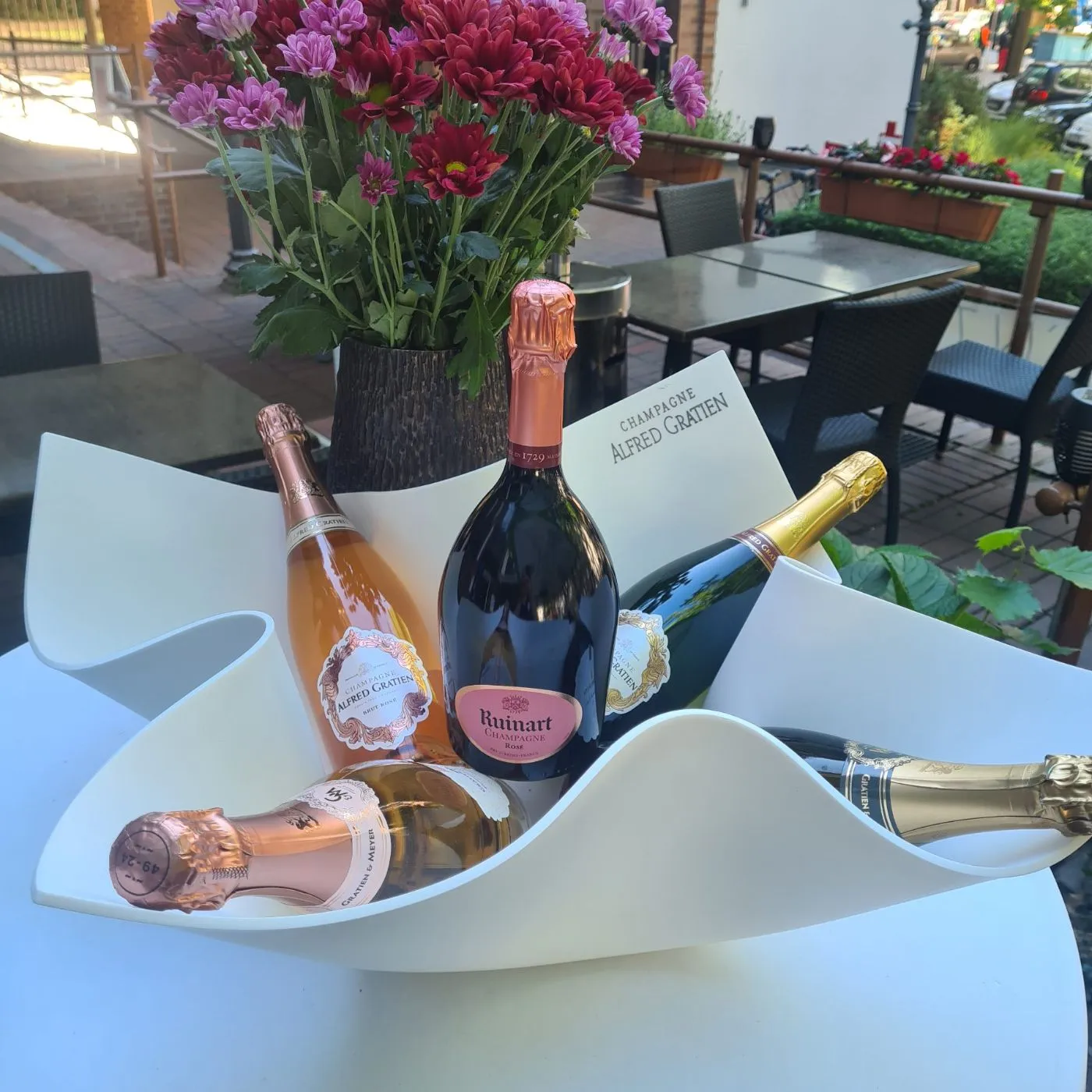 Mehrere Flaschen Champagner in einem geschwungenen weißen Kühler auf einem Terrassentisch, dahinter pinke Blumen.