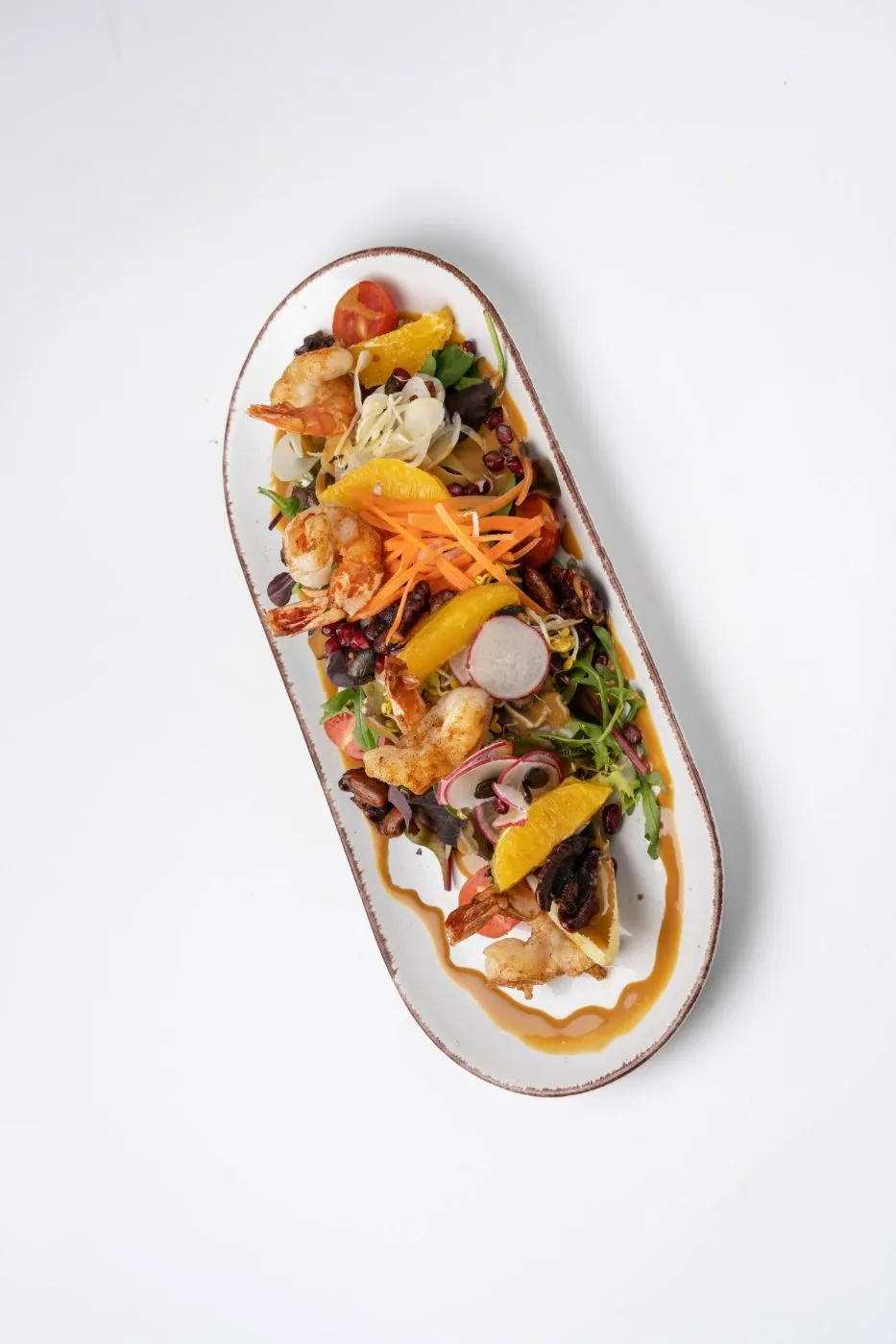 Bunter gemischter Salat mit gebratenen Garnelen, Orangenfilets, Granatapfel, Radieschen und Nüssen auf einer länglichen Platte, von oben.