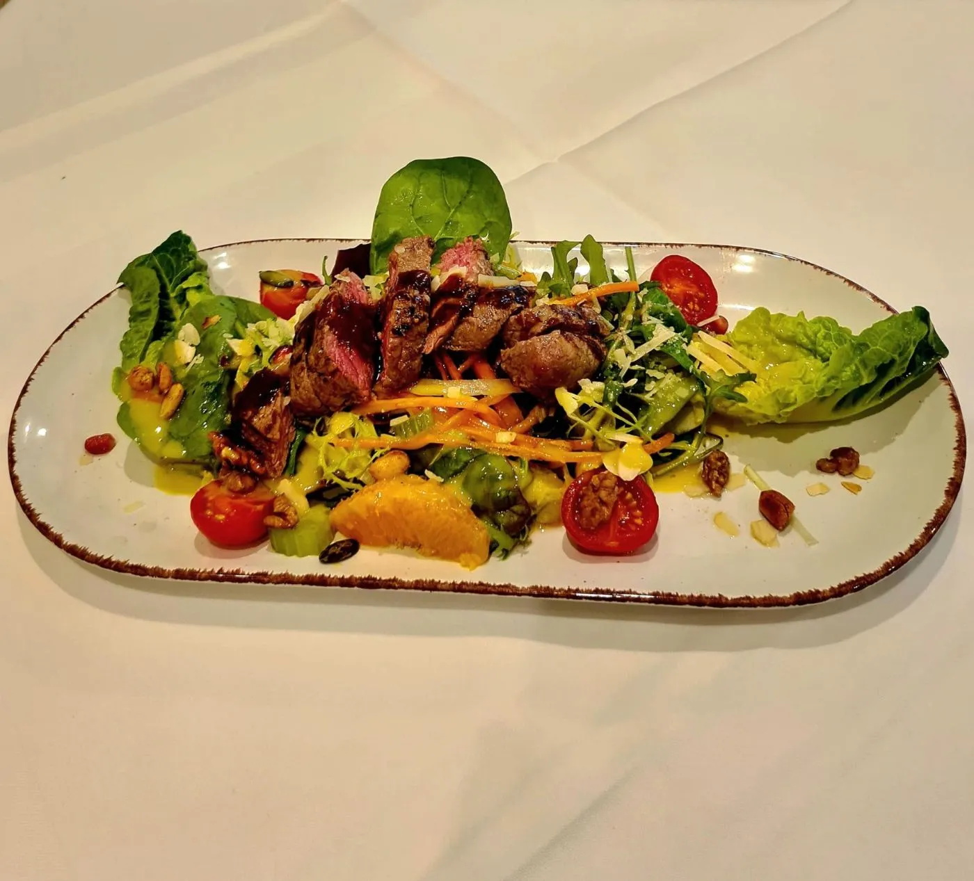 Salat mit gebratenen Rindfleischstreifen, Kirschtomaten und Orangen auf einer länglichen Platte auf weißer Tischdecke.