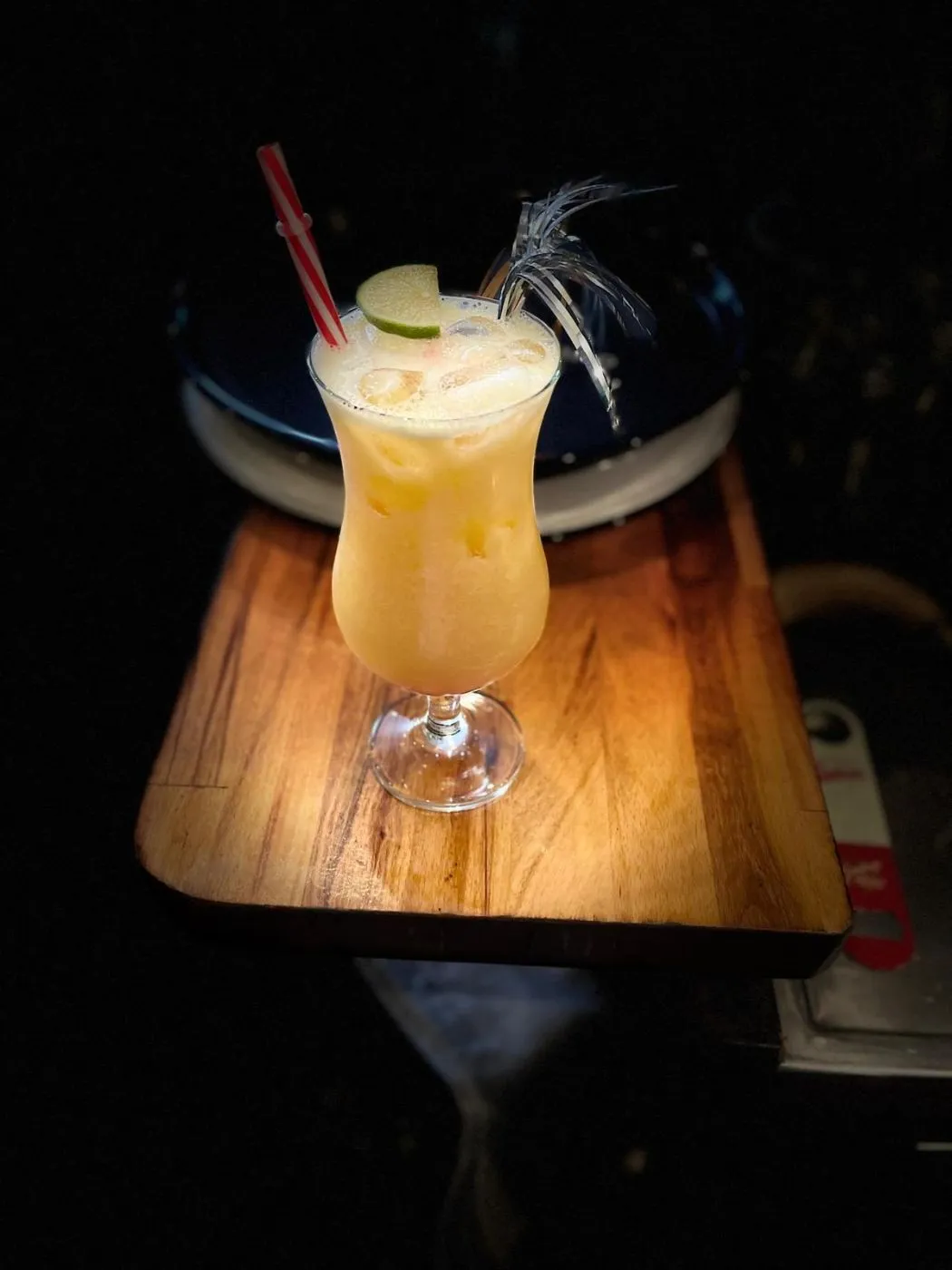 Heller Cocktail mit Strohhalm und Limette in einem geschwungenen Glas auf einem beleuchteten Holzbrett.