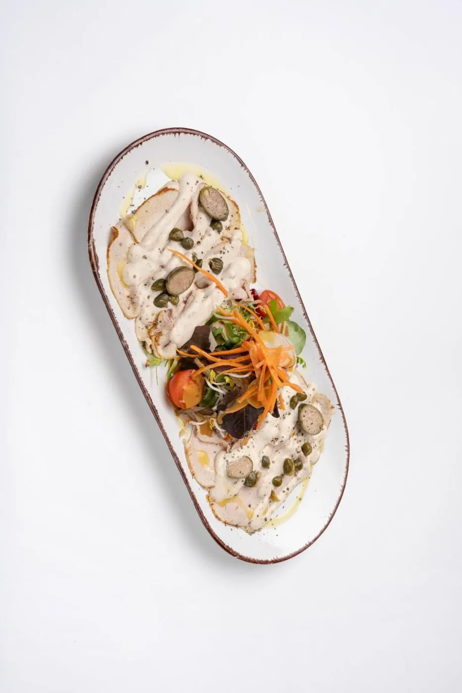 Vitello tonnato: rosa Kalbfleisch mit Thunfischcreme, Kapernäpfeln und Salatgarnitur auf einem ovalen Teller, von oben.