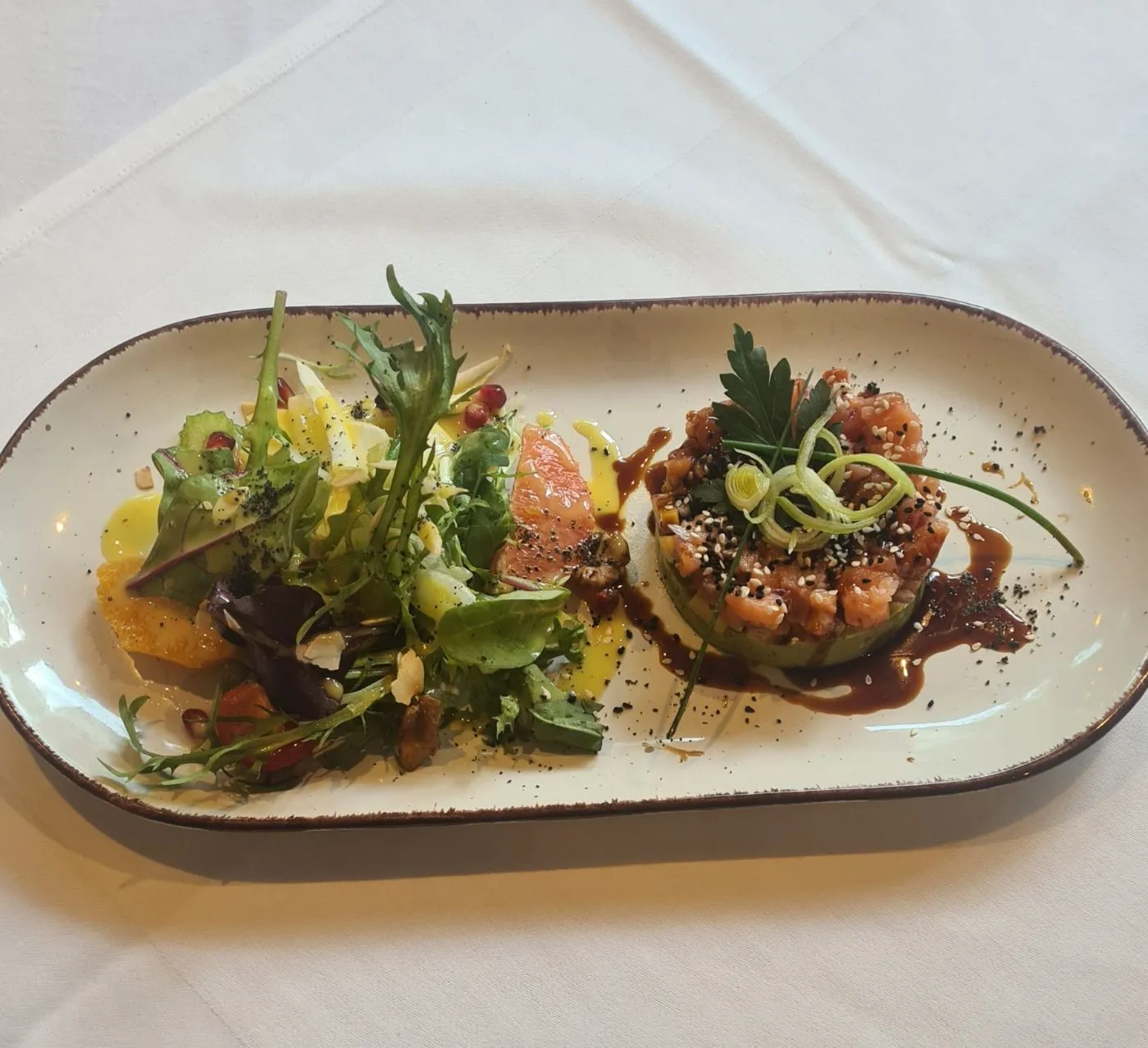 Lachstatar-Türmchen auf Avocado mit Sesam und Teriyaki, daneben Blattsalat mit Orange und Granatapfel auf einer länglichen Platte.