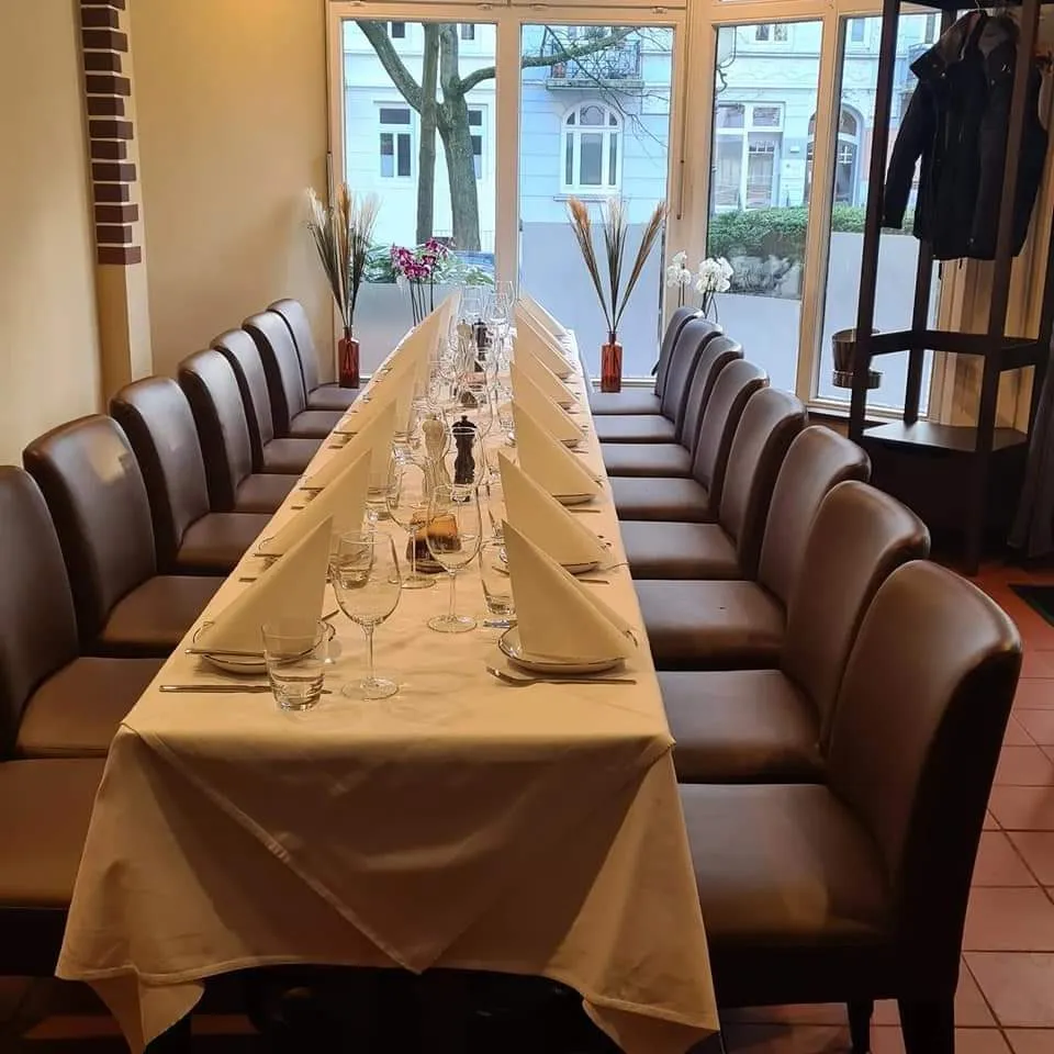 Lange Tafel mit beiger Tischdecke, gefalteten Servietten und Weingläsern vor der Fensterfront.