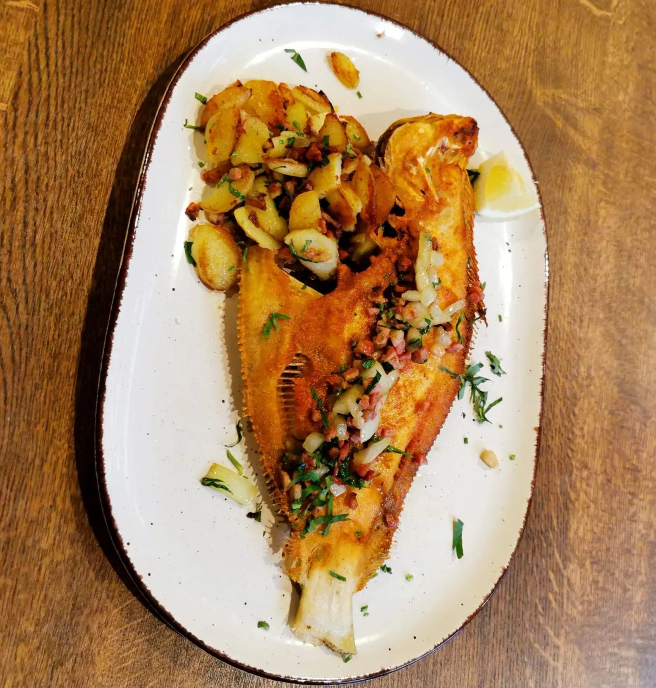 Gebratenes Fischfilet mit Bratkartoffeln und Kräutern auf einem ovalen Teller, auf einem Holztisch.